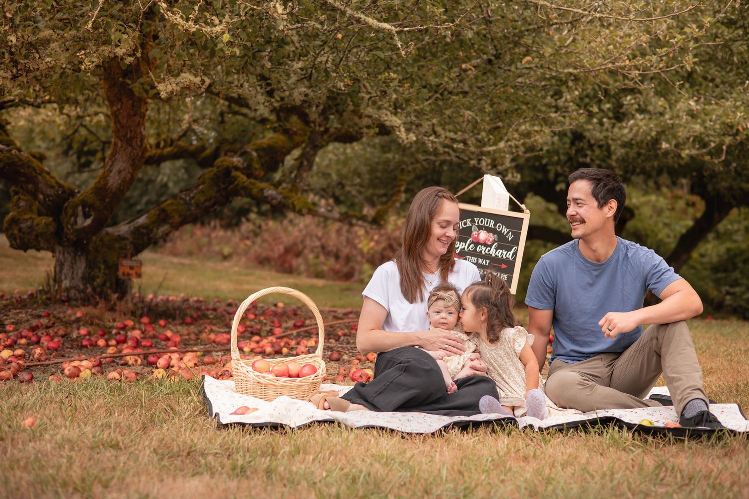 Family picnic under apple trees with basket of red apples, blanket, and chalkboard sign in a scenic orchard.