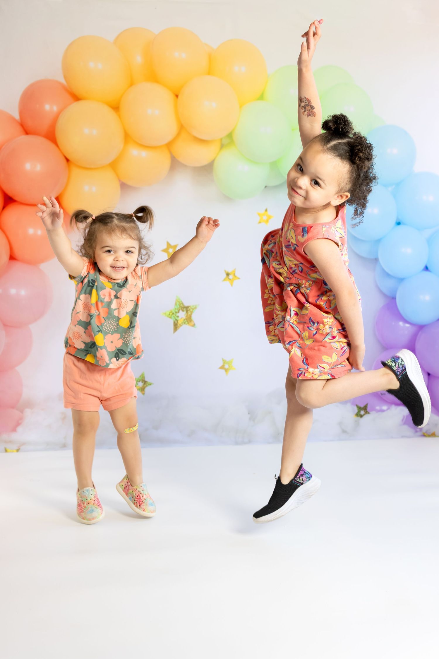 Two joyful girls in floral outfits jumping in front of a colorful rainbow balloon arch with gold stars.