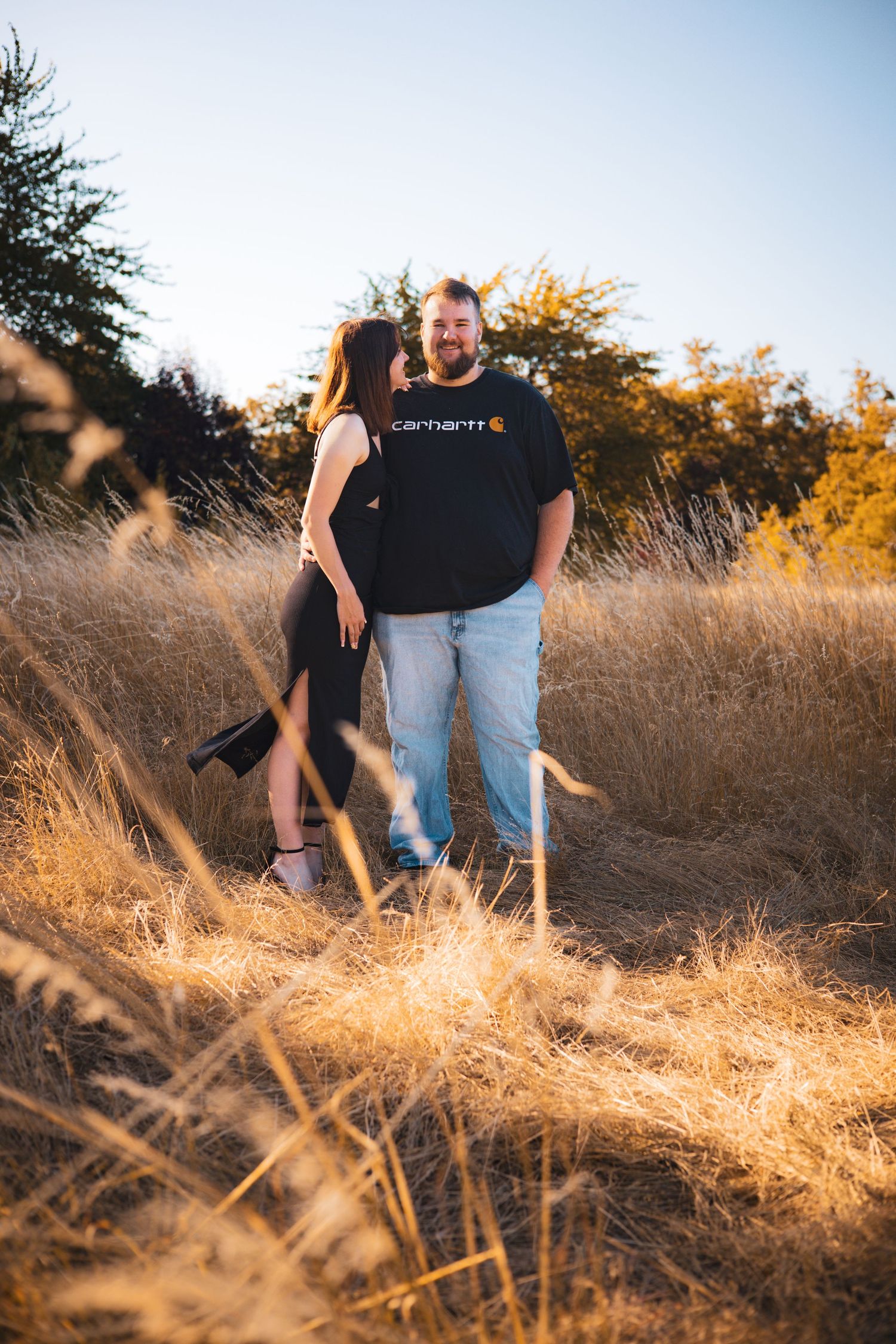 Couple stands together in tall golden grass during outdoor sunset session.