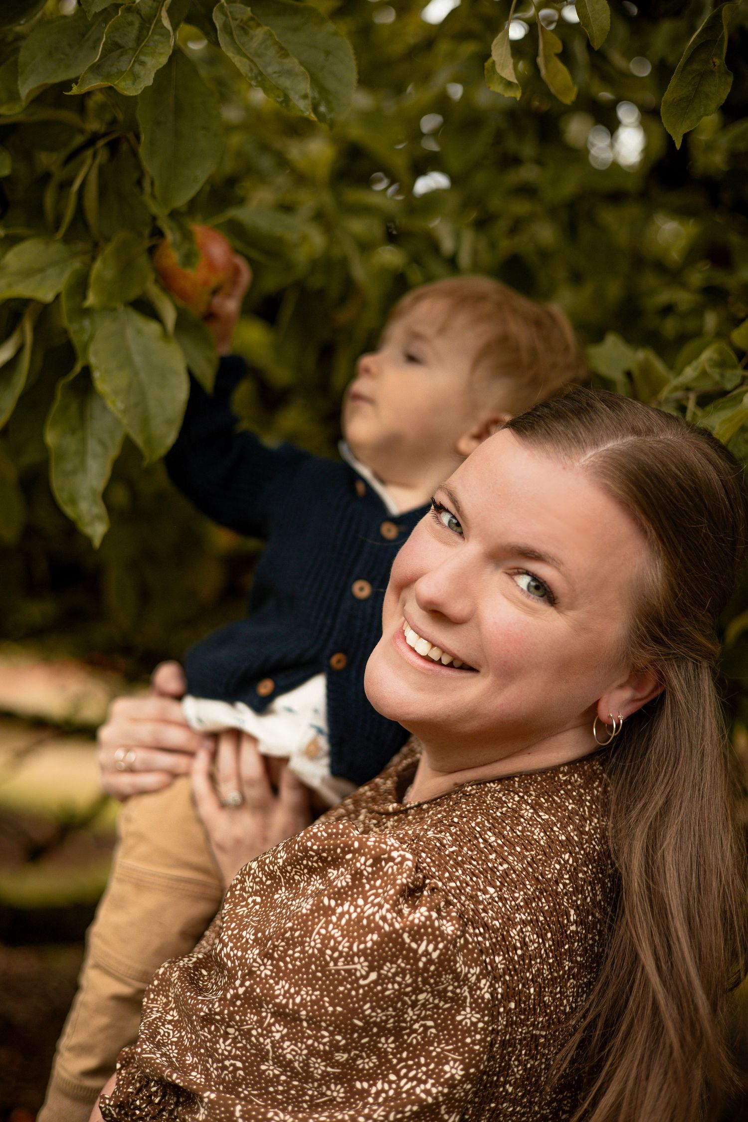 Smiling woman in brown sequin top holds child reaching for apple in lush green orchard tree.