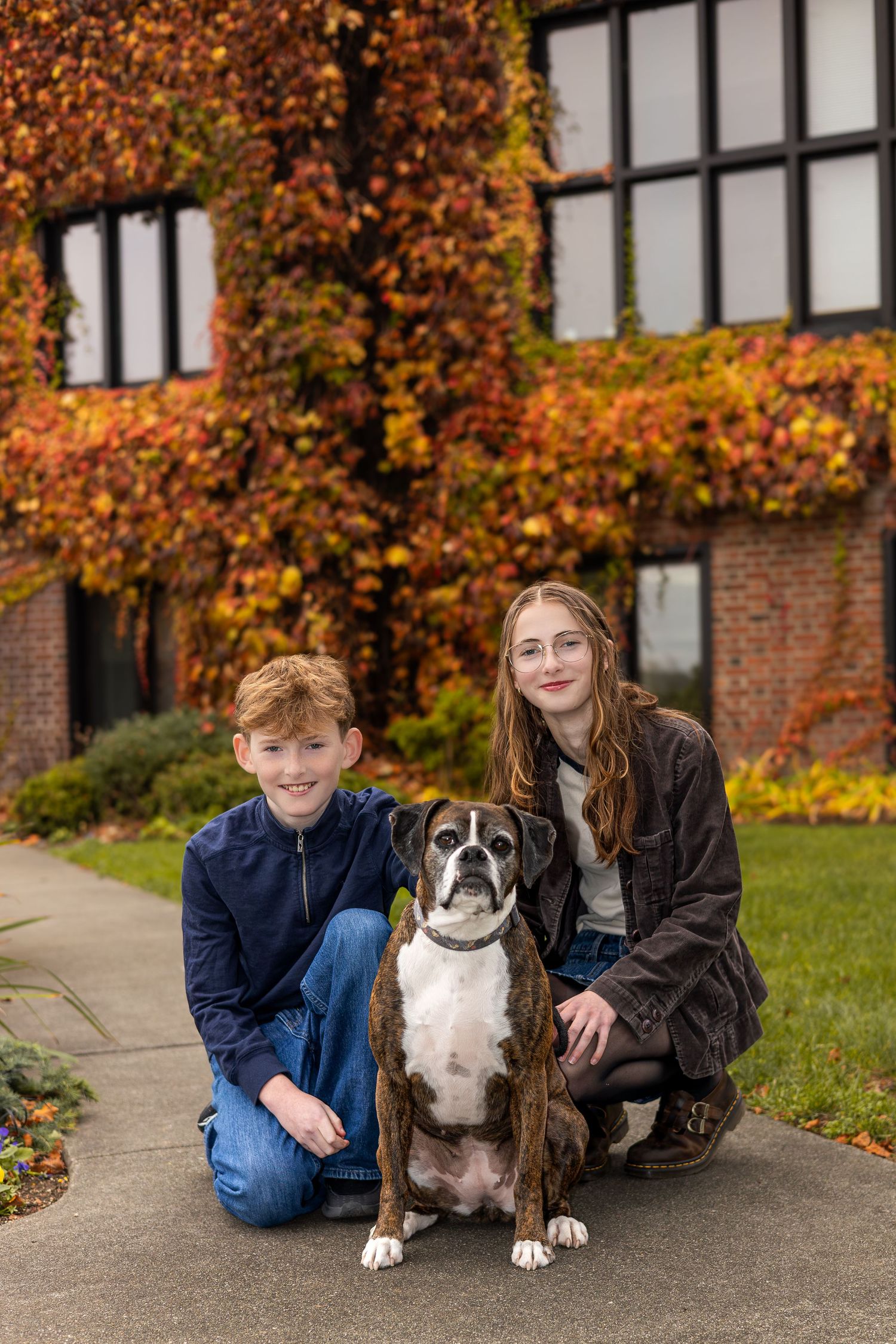 Two kids and a Boxer dog pose outdoors in front of a brick building covered in colorful autumn ivy foliage.