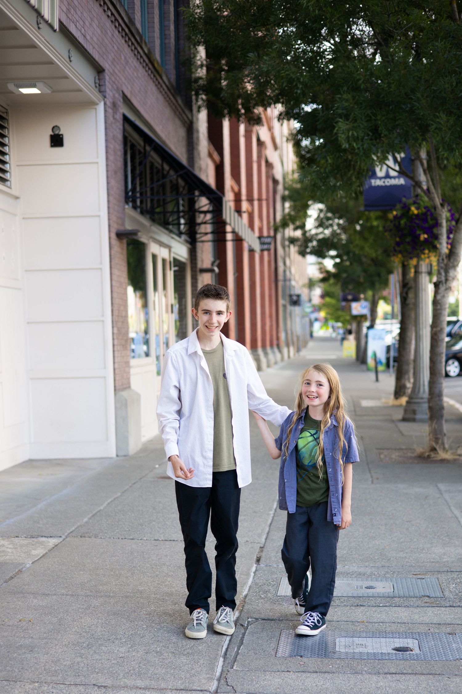 Two people walking together on a city sidewalk with brick buildings and trees in the background.