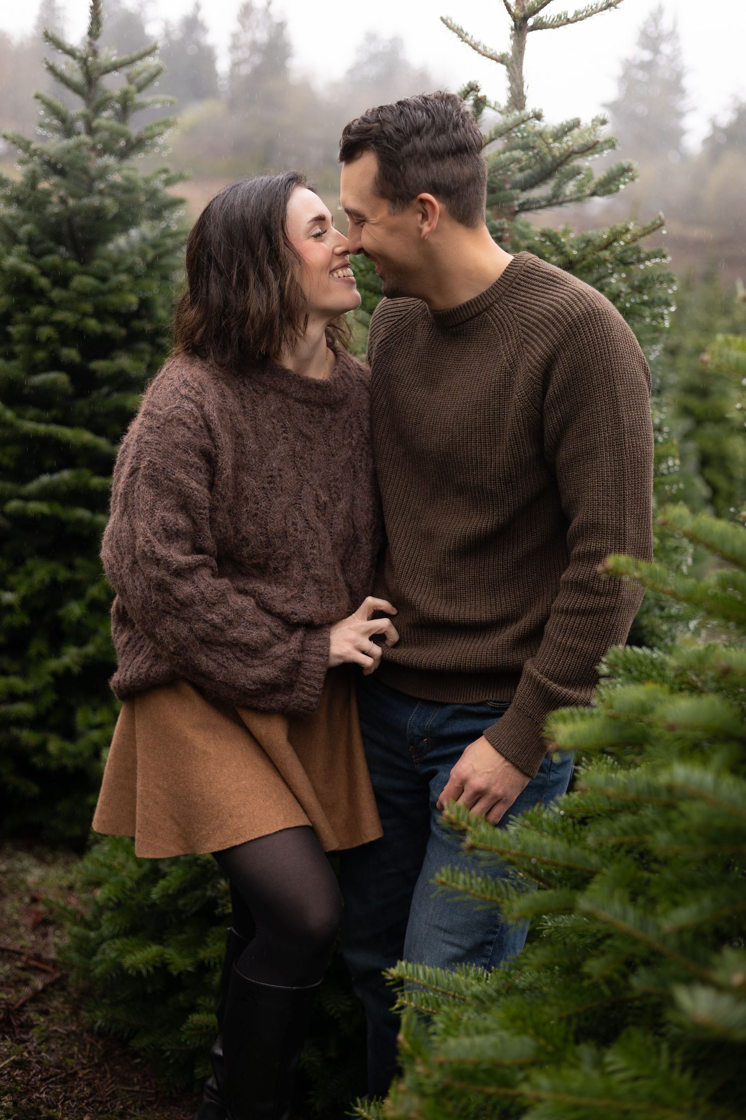 A couple in cozy brown sweaters share an intimate moment among evergreen trees at a Christmas tree farm.