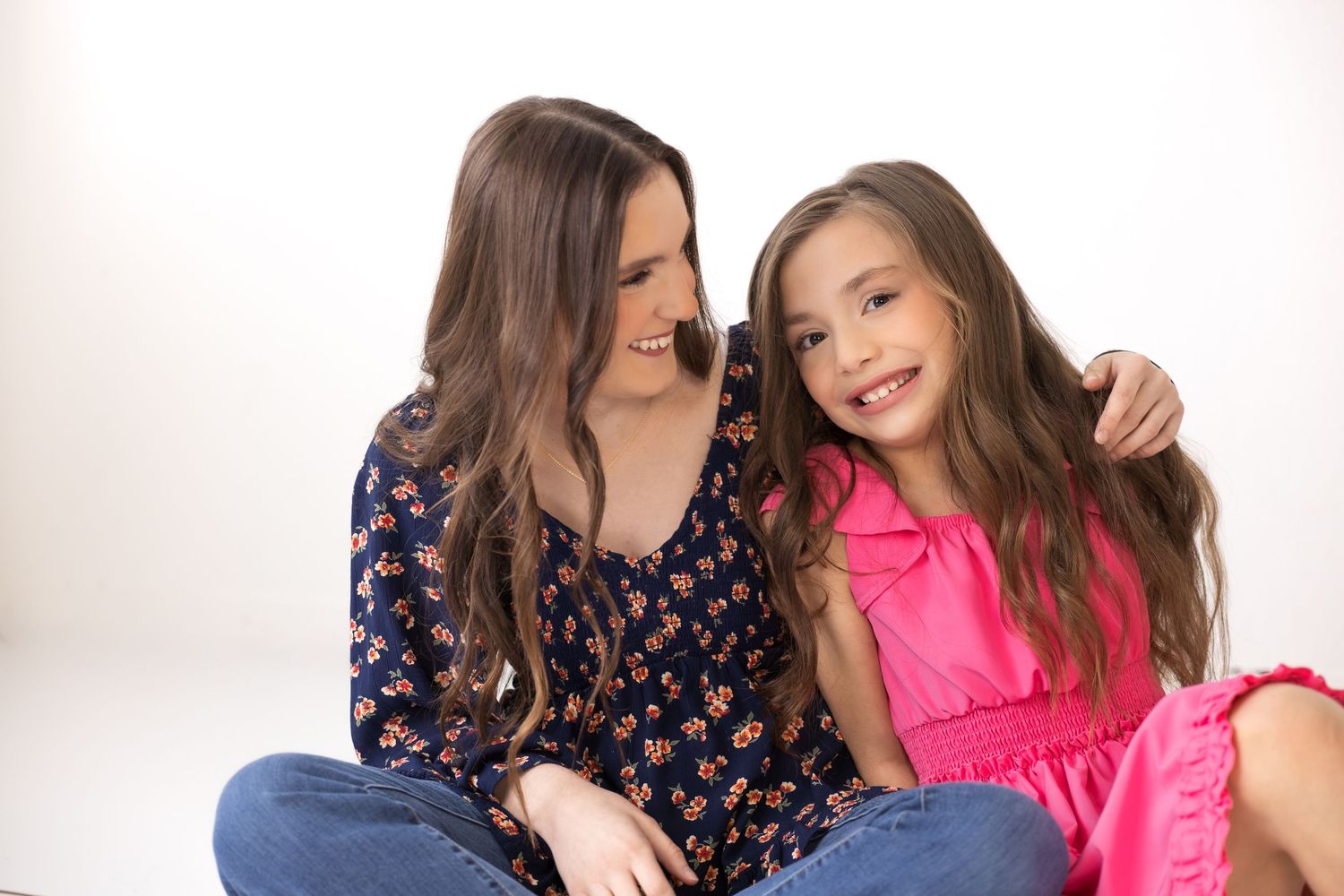 A woman and young girl with long brown hair smile together, the woman's arm around the girl, against a white background.