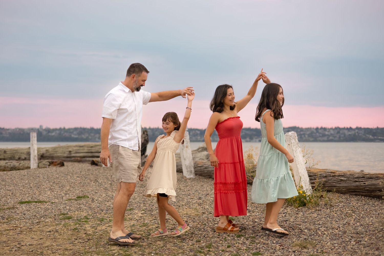 Family of four dancing on a rocky beach at sunset, wearing summer dresses and casual attire.