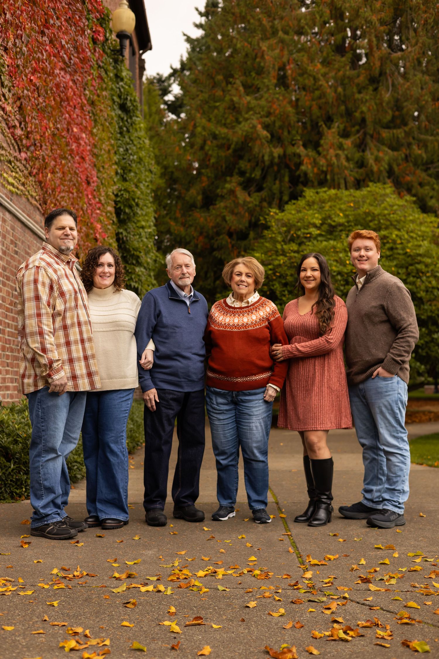 Family of six posing outdoors in fall, wearing cozy sweaters and jeans, surrounded by colorful autumn leaves and greenery.