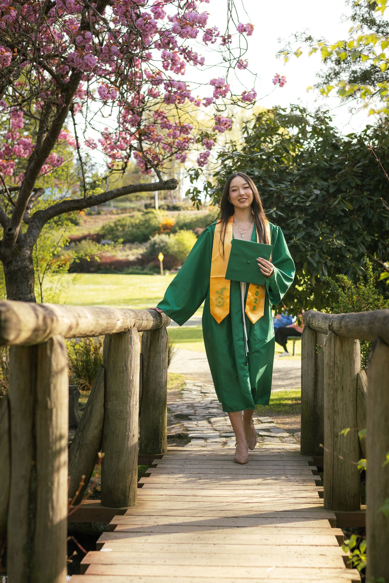 Graduate in green cap and gown poses on wooden bridge under blooming cherry blossoms during spring photo session.
