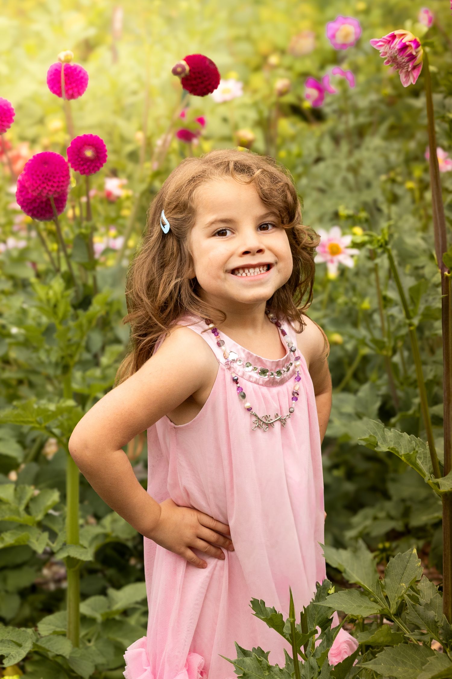 Smiling little girl in pink dress posing among colorful dahlia flowers in a garden.