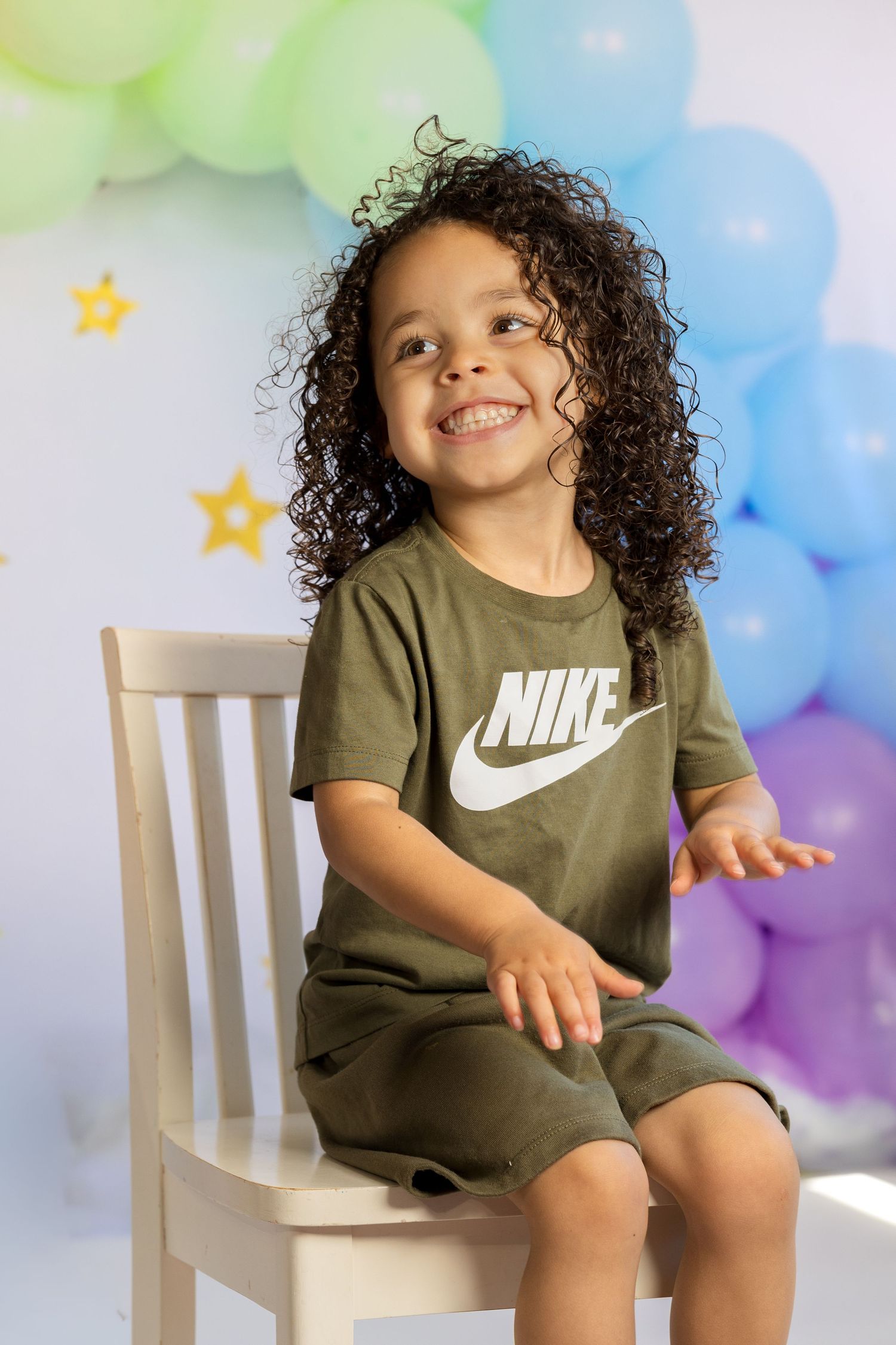 Smiling child with curly hair wearing olive Nike outfit sits on white chair with colorful balloons and stars backdrop.