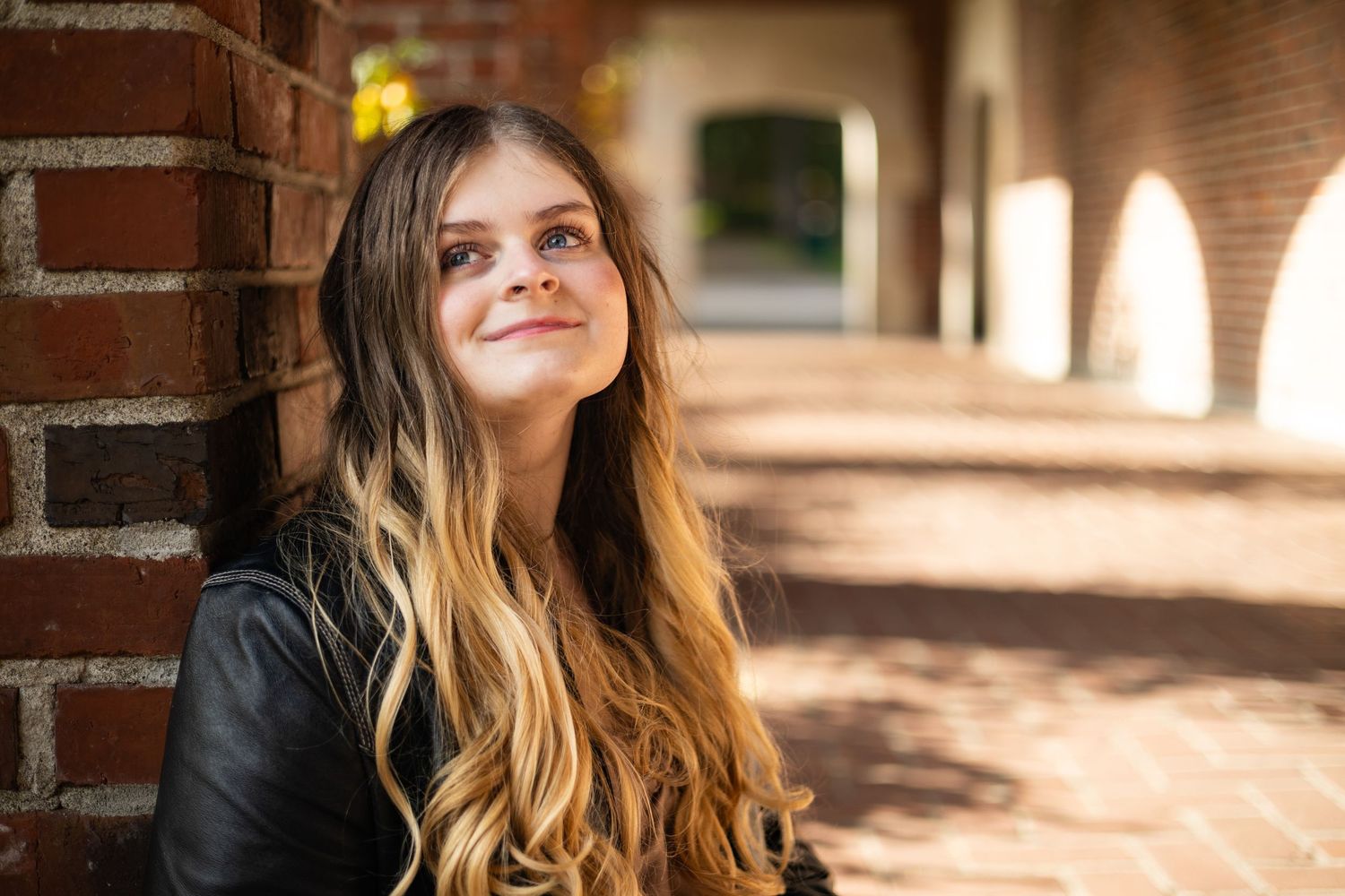 Young woman with long ombre hair smiling outdoors near brick wall in soft natural light.