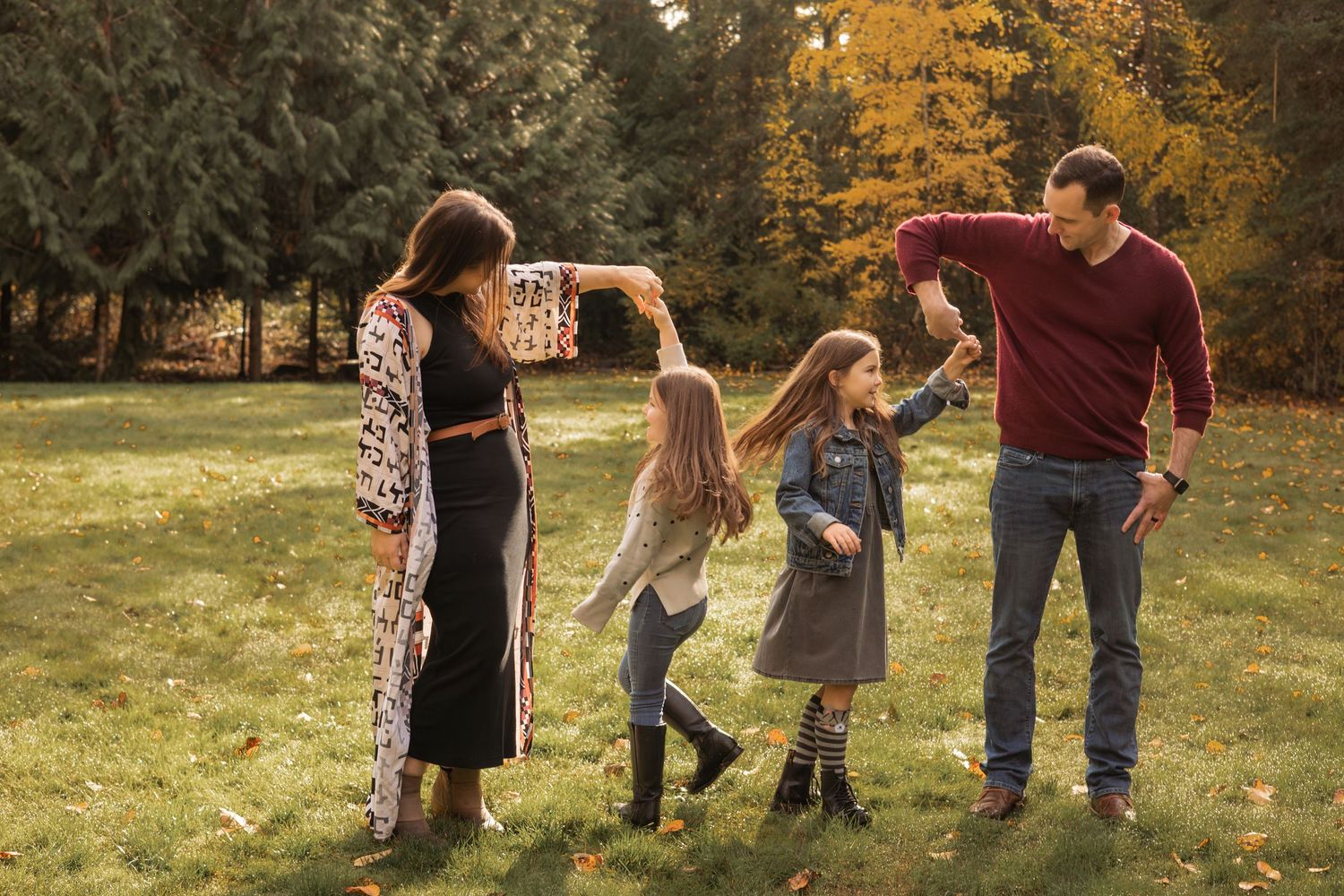 Family of four dancing outdoors in autumn park, parents spinning two girls among colorful fall foliage.