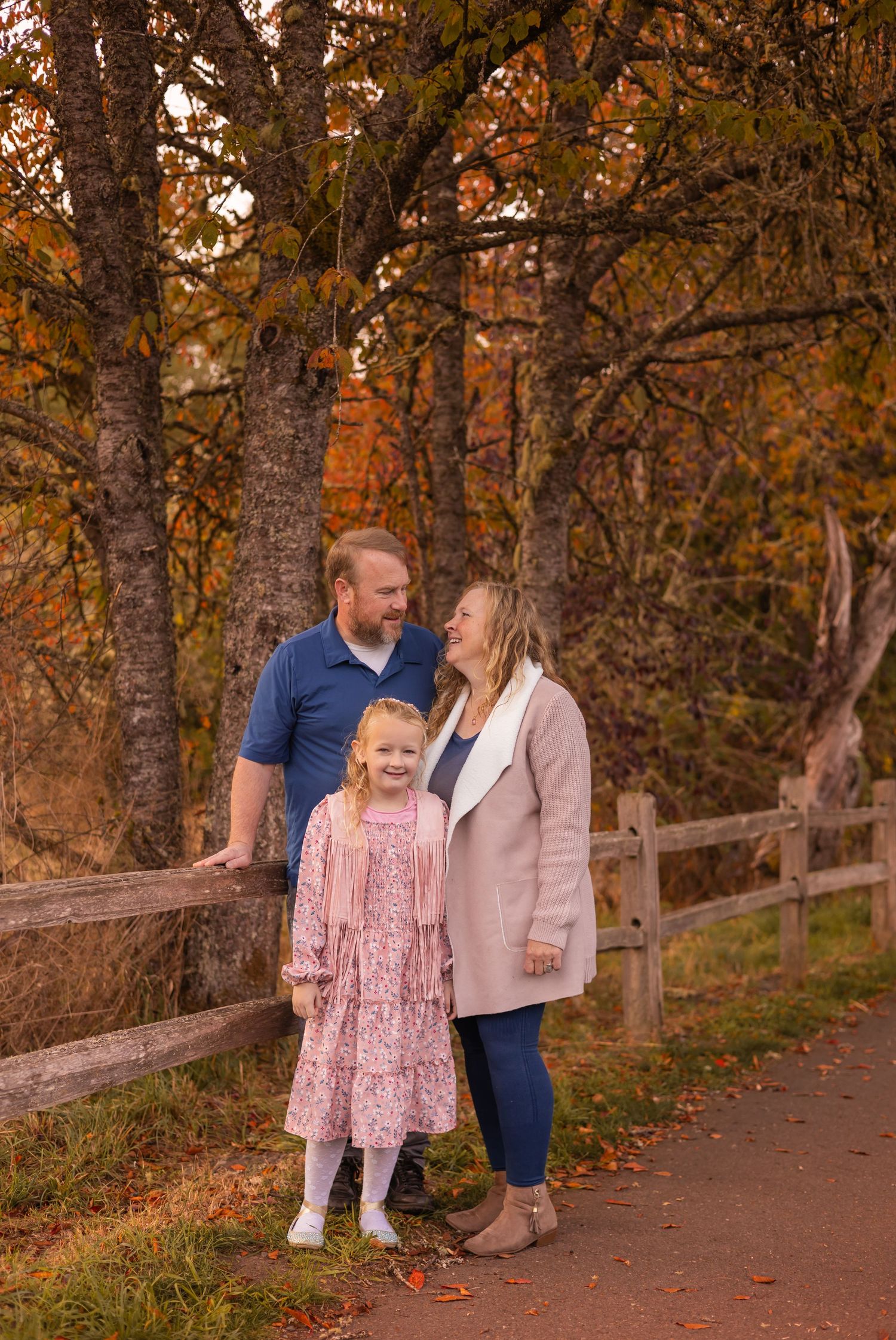 Family of three posing by a wooden fence surrounded by vibrant autumn foliage on a scenic fall day.