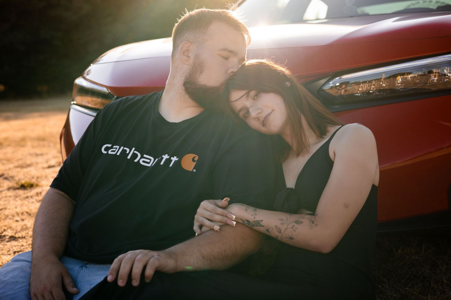 Couple shares tender moments next to red sports car during outdoor photoshoot.