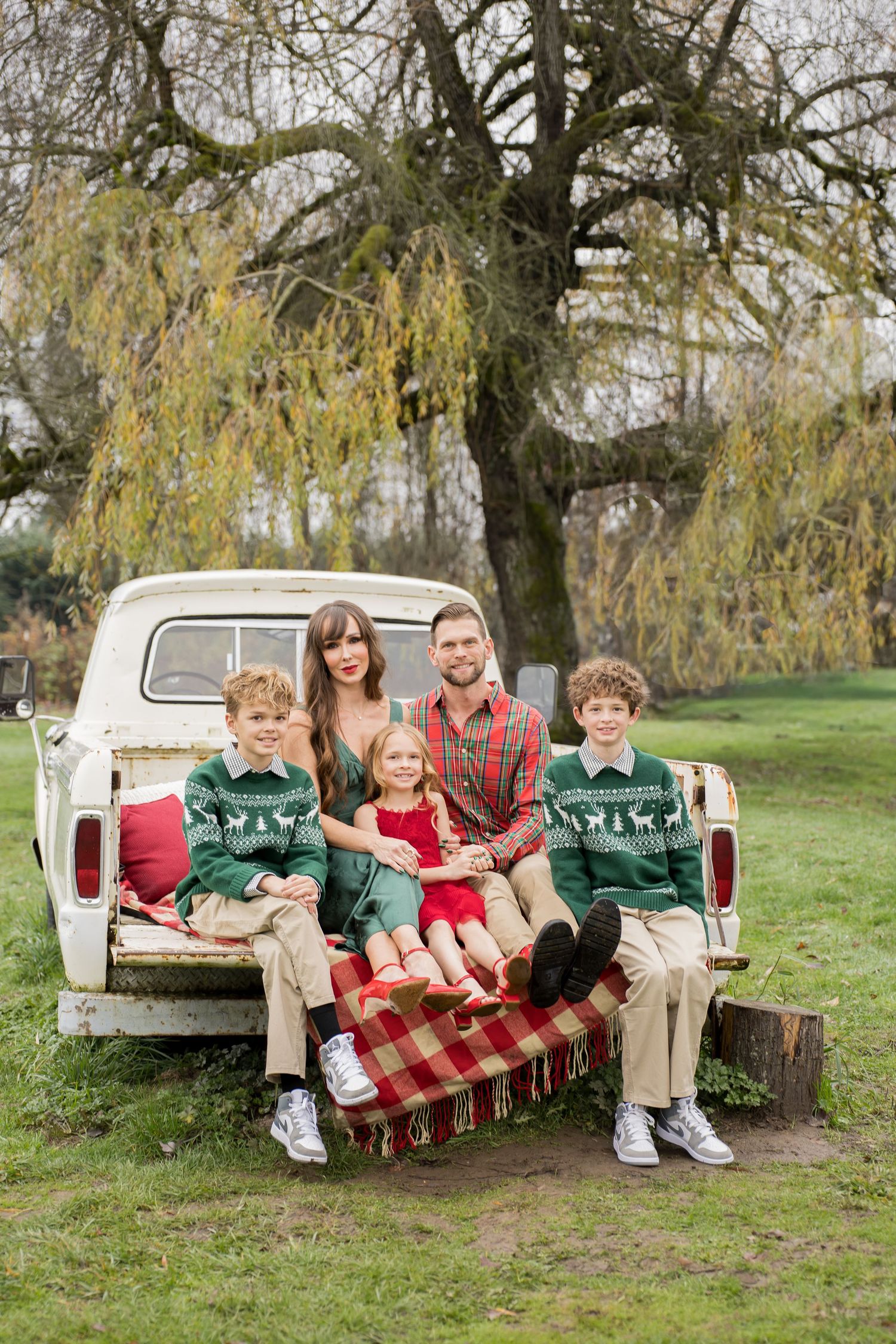 A family sits together on a vintage white pickup truck tailgate with a red plaid blanket on a grassy hillside.