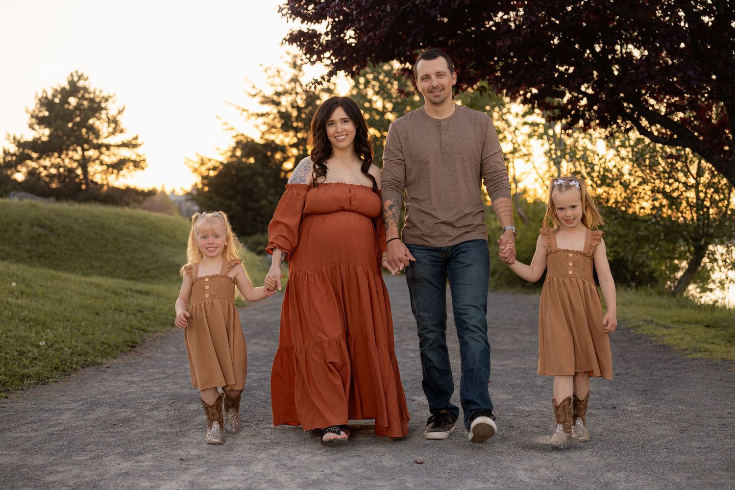 Pregnant woman in rust dress walks with husband and two daughters on a path during golden hour family photo session.