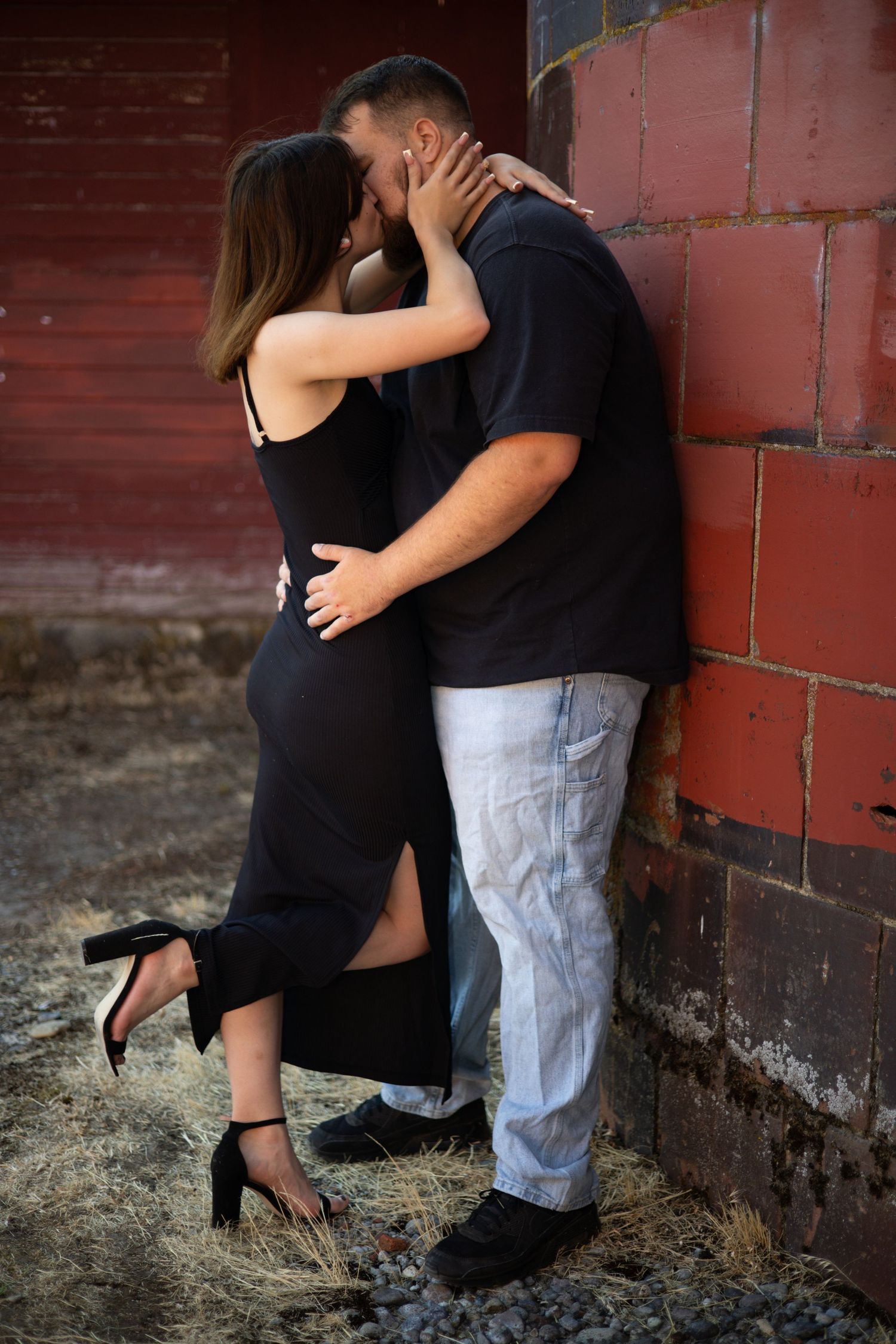 A romantic couple embraces against a rustic brick wall, with the woman wearing a black dress and heels.