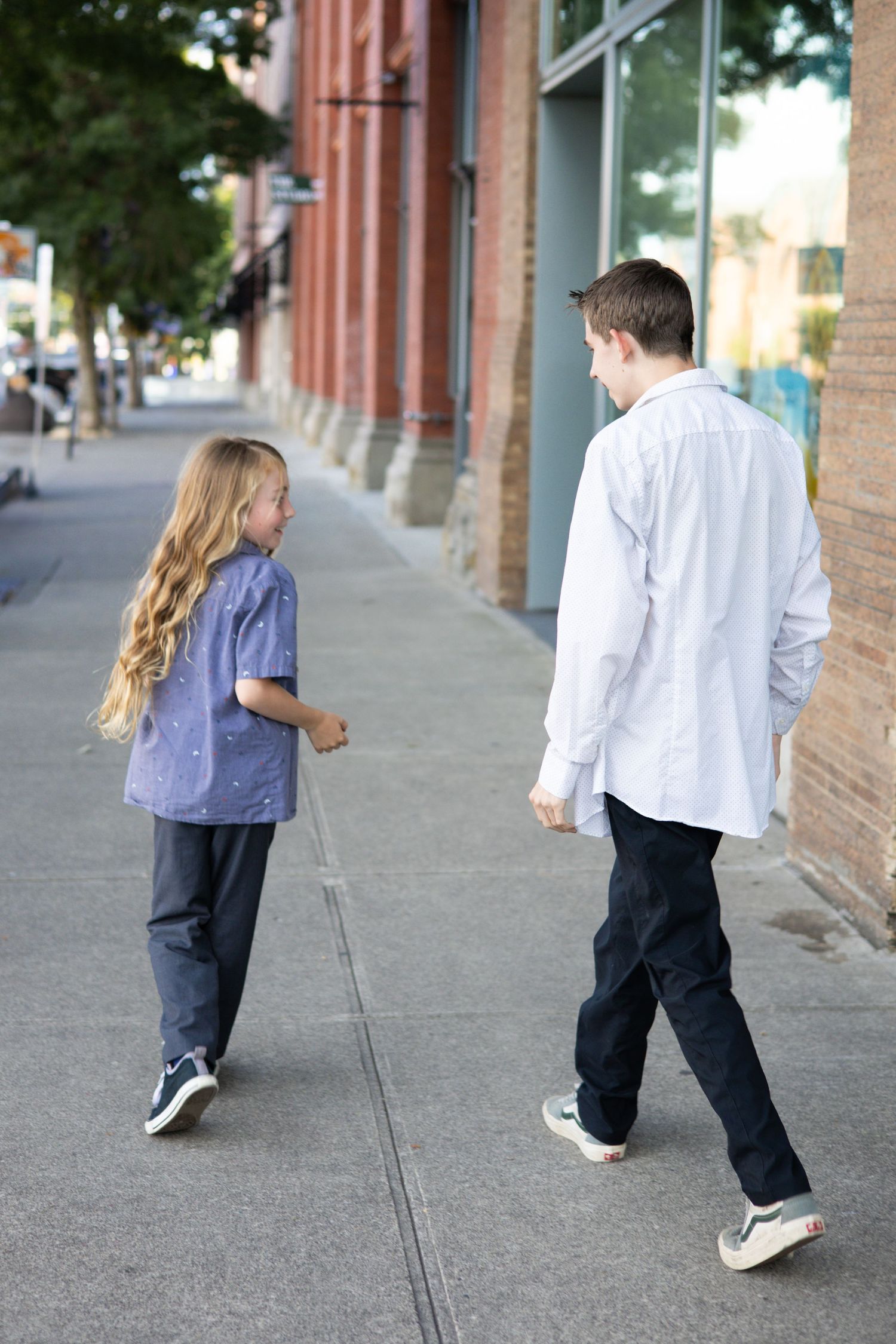 People walking along an urban sidewalk with brick buildings and trees visible.