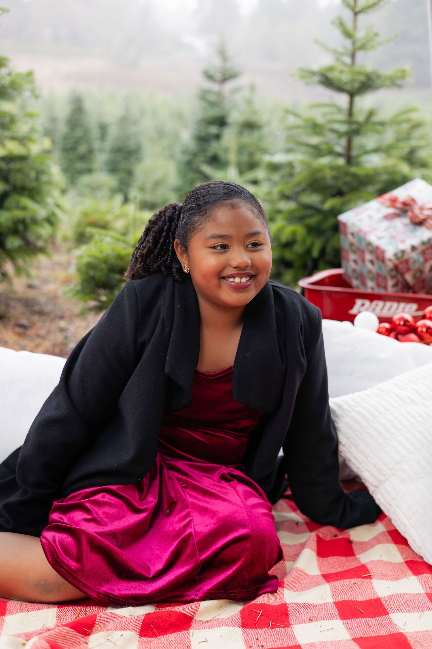 A person in a black blazer and magenta dress poses on a red and white checkered blanket outdoors among evergreen trees.