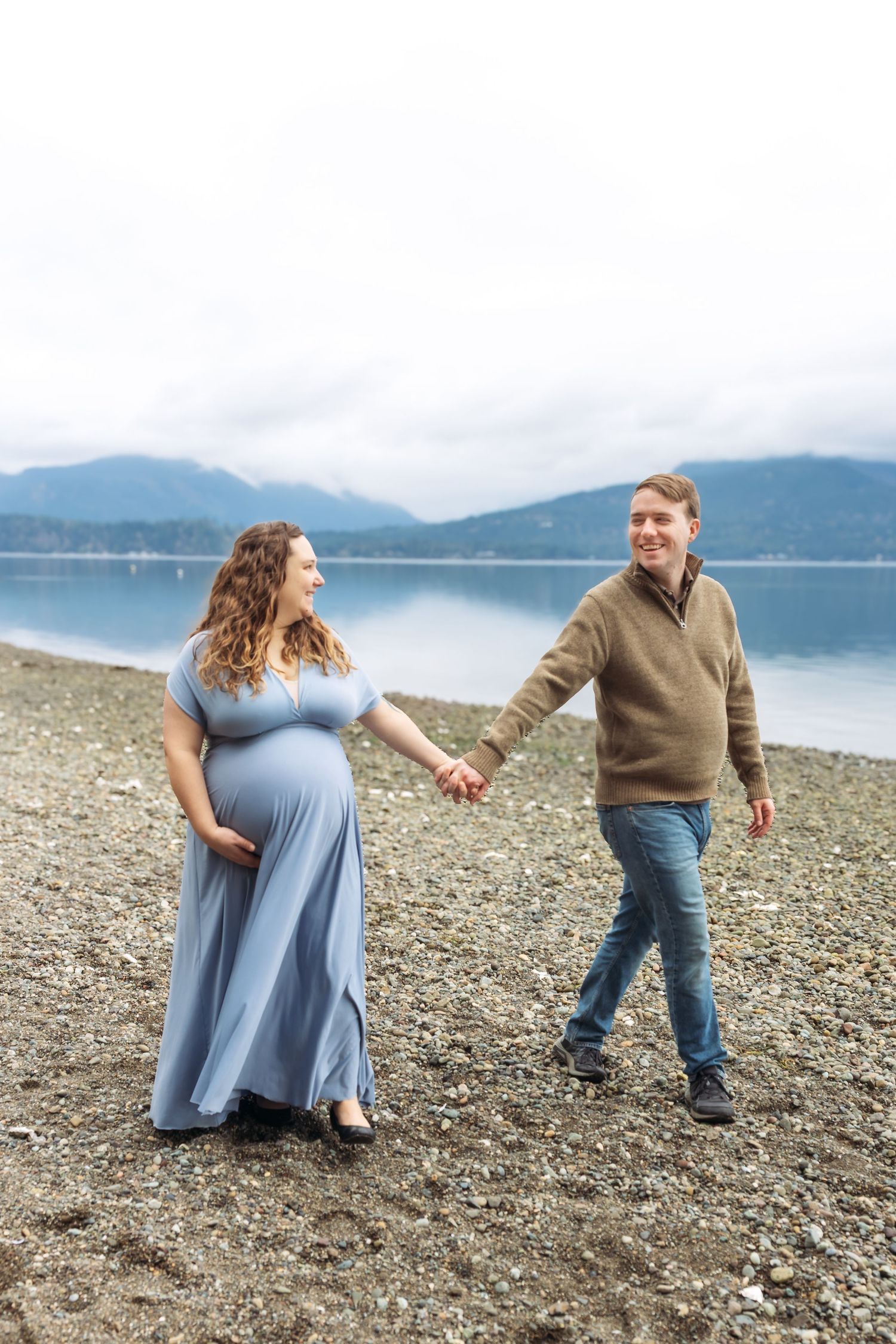 Pregnant couple holding hands and walking on a rocky beach with mountains and calm water in the background.