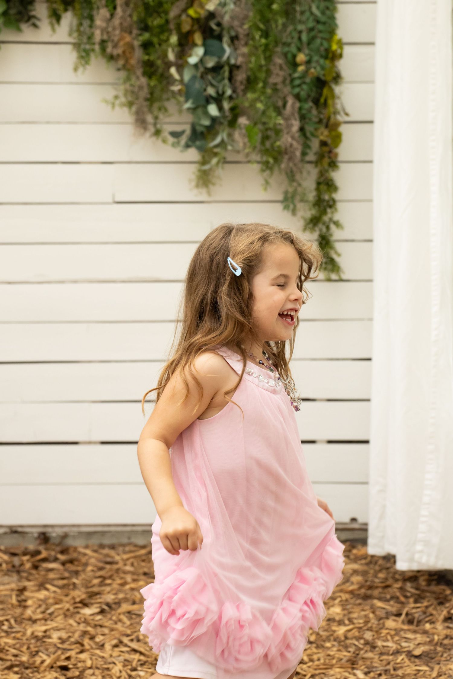 Young girl in pink ruffled dress laughing and twirling outdoors near white wall with hanging greenery.