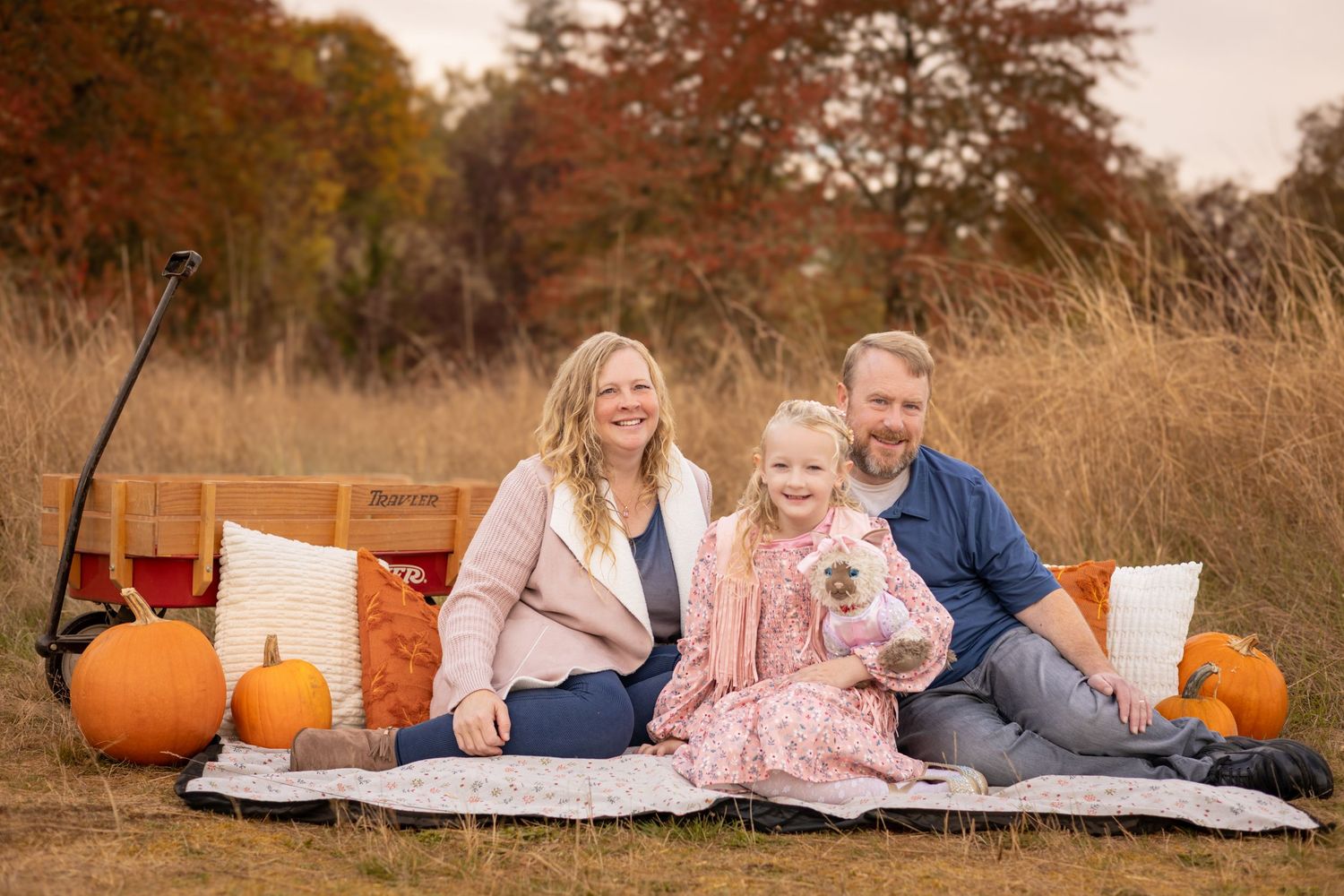 Family of three sitting on a blanket in an autumn field surrounded by pumpkins for a fall photo session.