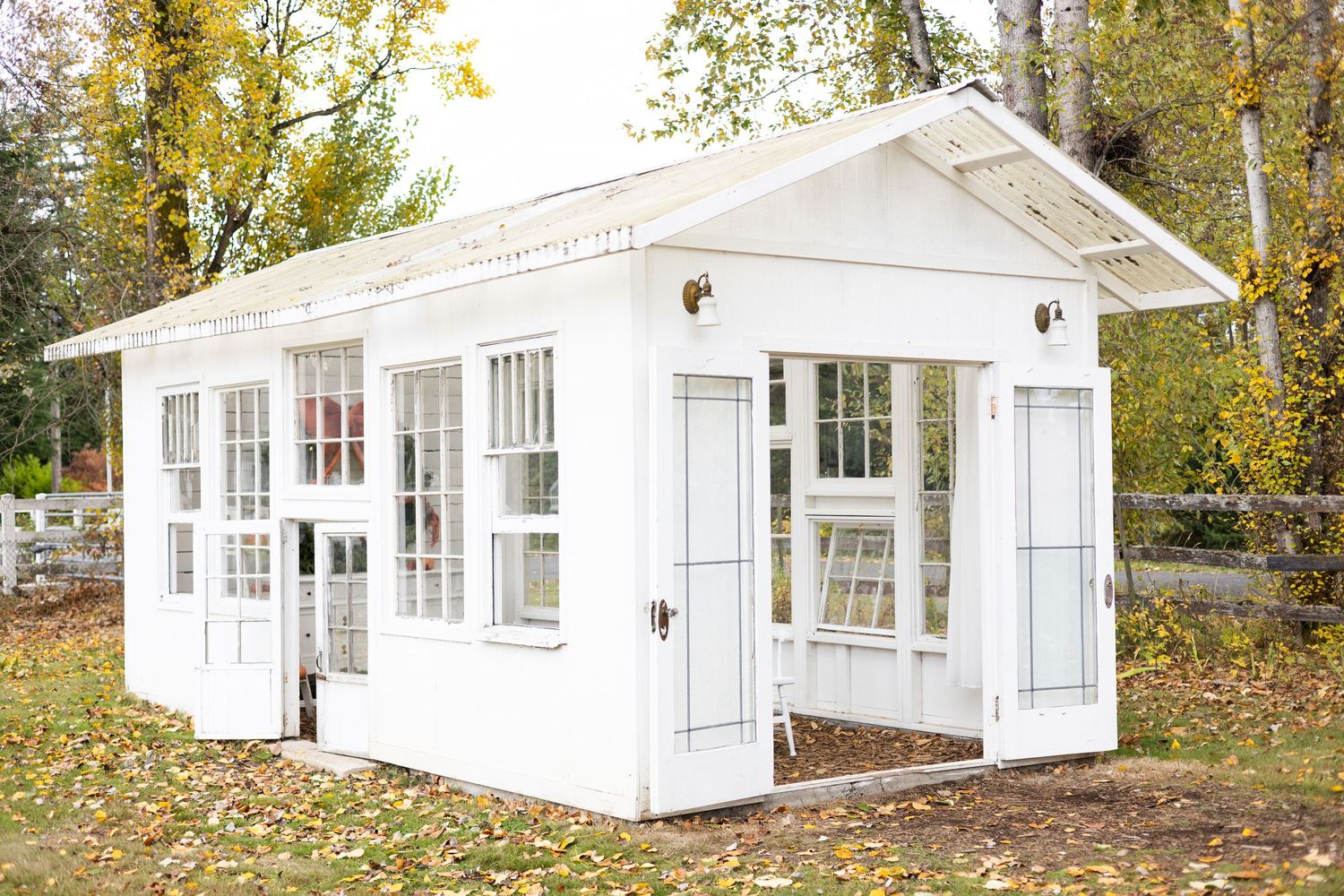 A white summer cottage with multiple windows and glass doors sits nestled among autumn trees in a wooded setting.