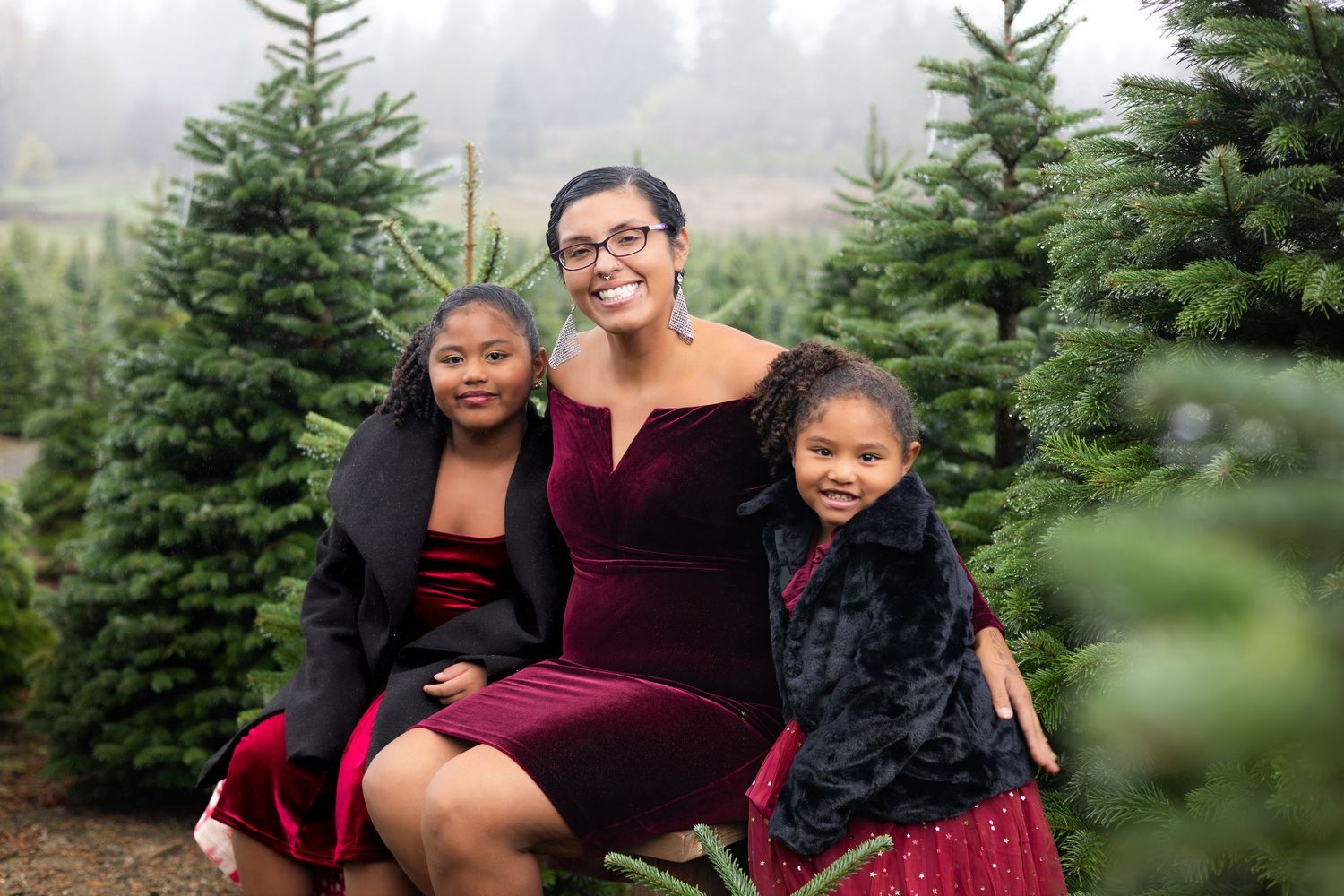 A family poses together among evergreen trees at a Christmas tree farm wearing festive burgundy and black outfits.
