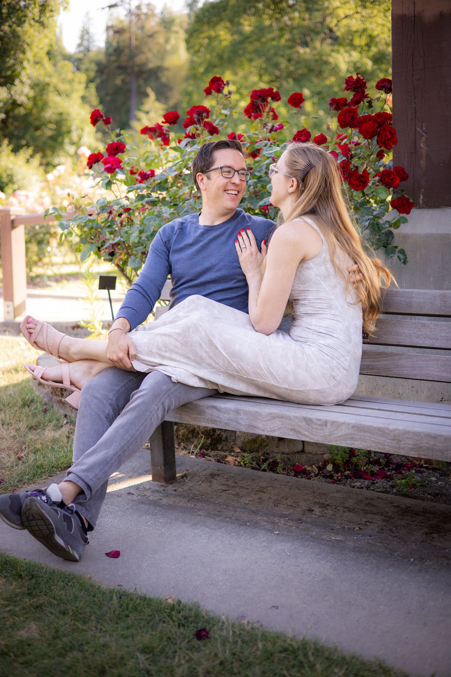 A couple relaxes together on a wooden bench near blooming red roses in a garden setting.