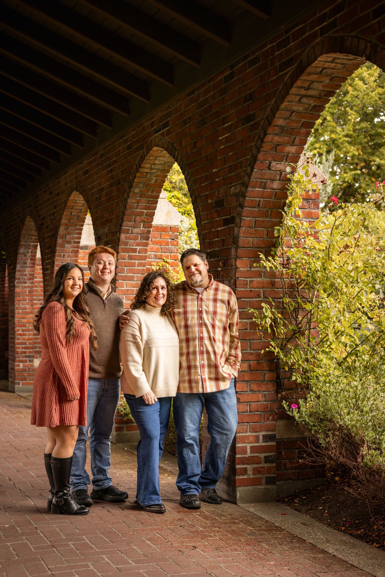 Family of four smiling under brick arches in autumn, wearing casual fall outfits of earth tones and jeans.