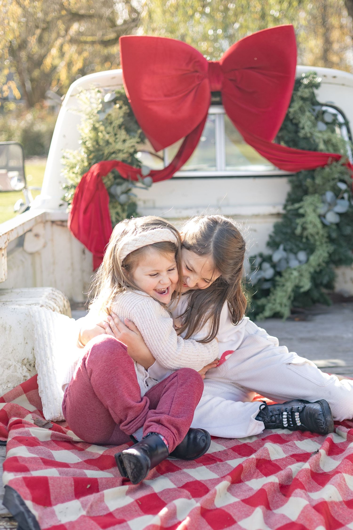 Two children share a sweet moment while laying on a red and white checkered blanket outdoors during a holiday photoshoot.