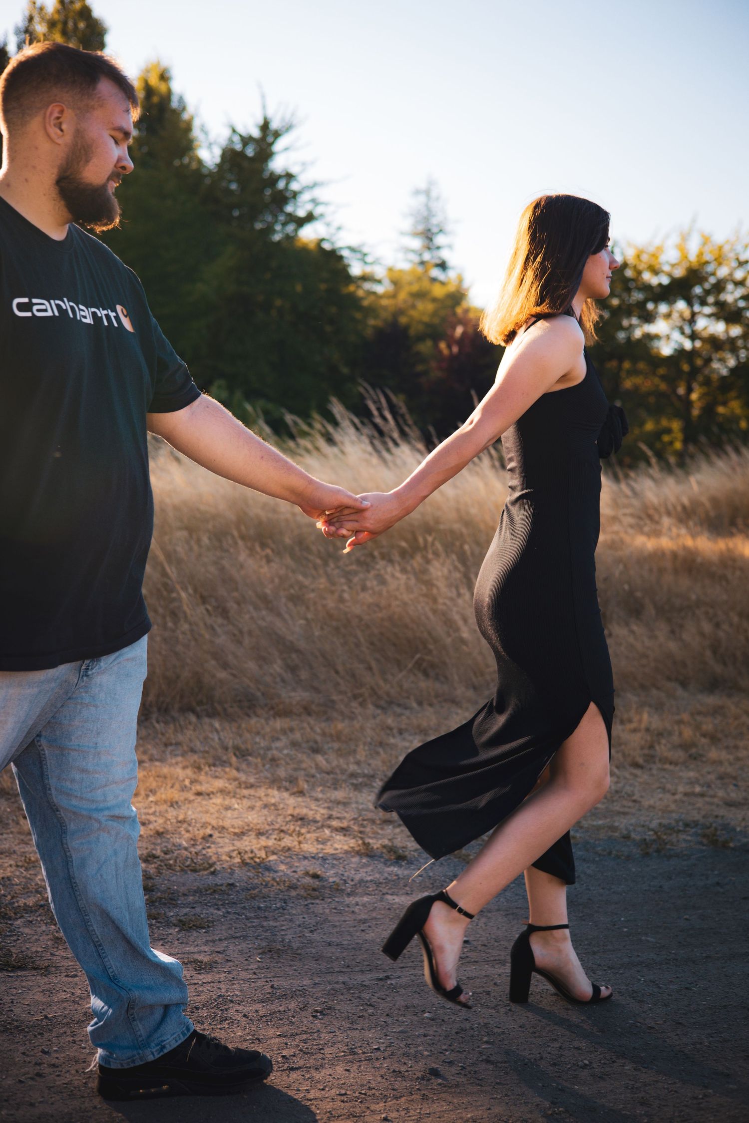 Couple holding hands in golden field at sunset with dramatic lighting and flowing black dress.