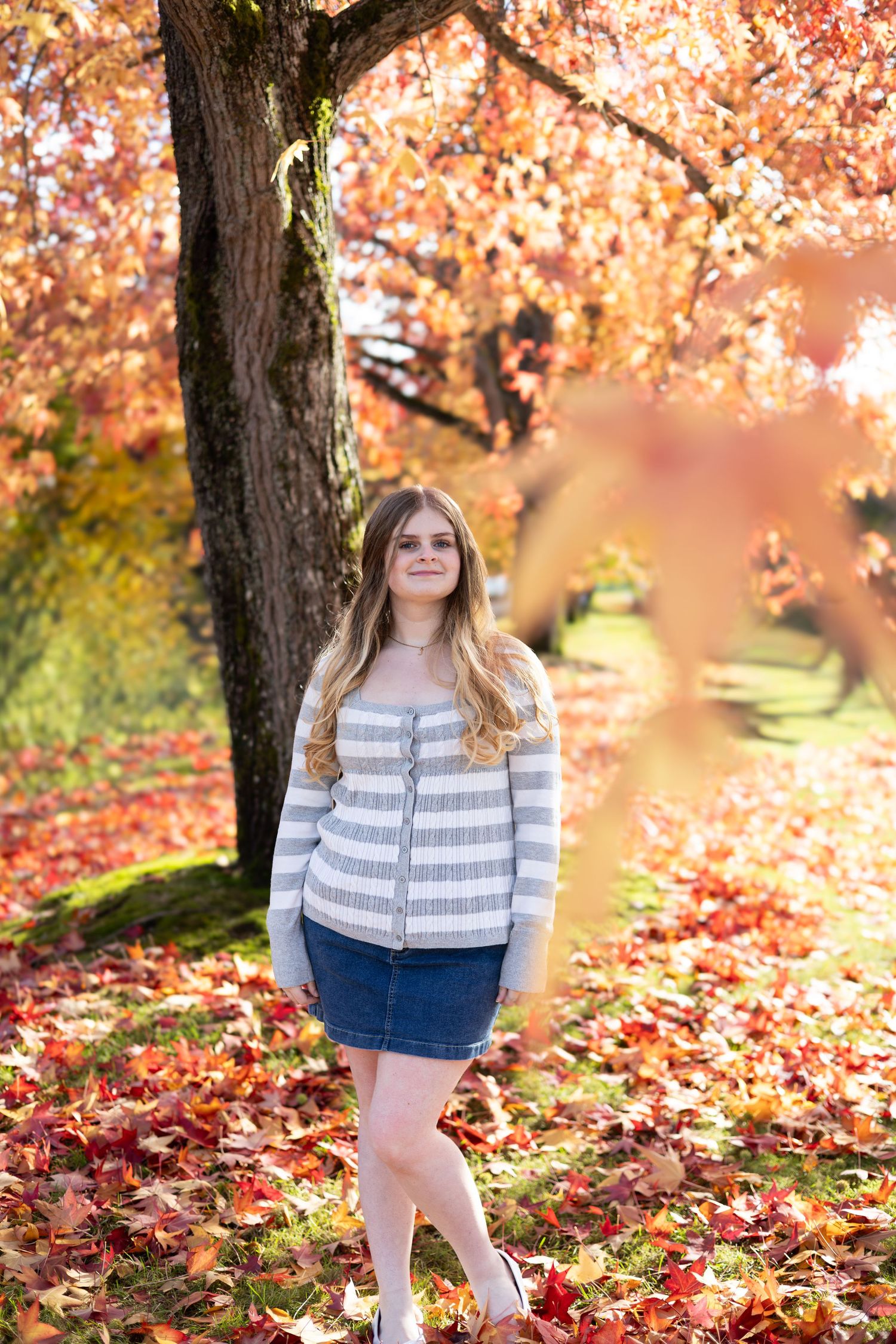 Young woman in striped sweater and denim skirt standing among vibrant red autumn leaves in a scenic park.
