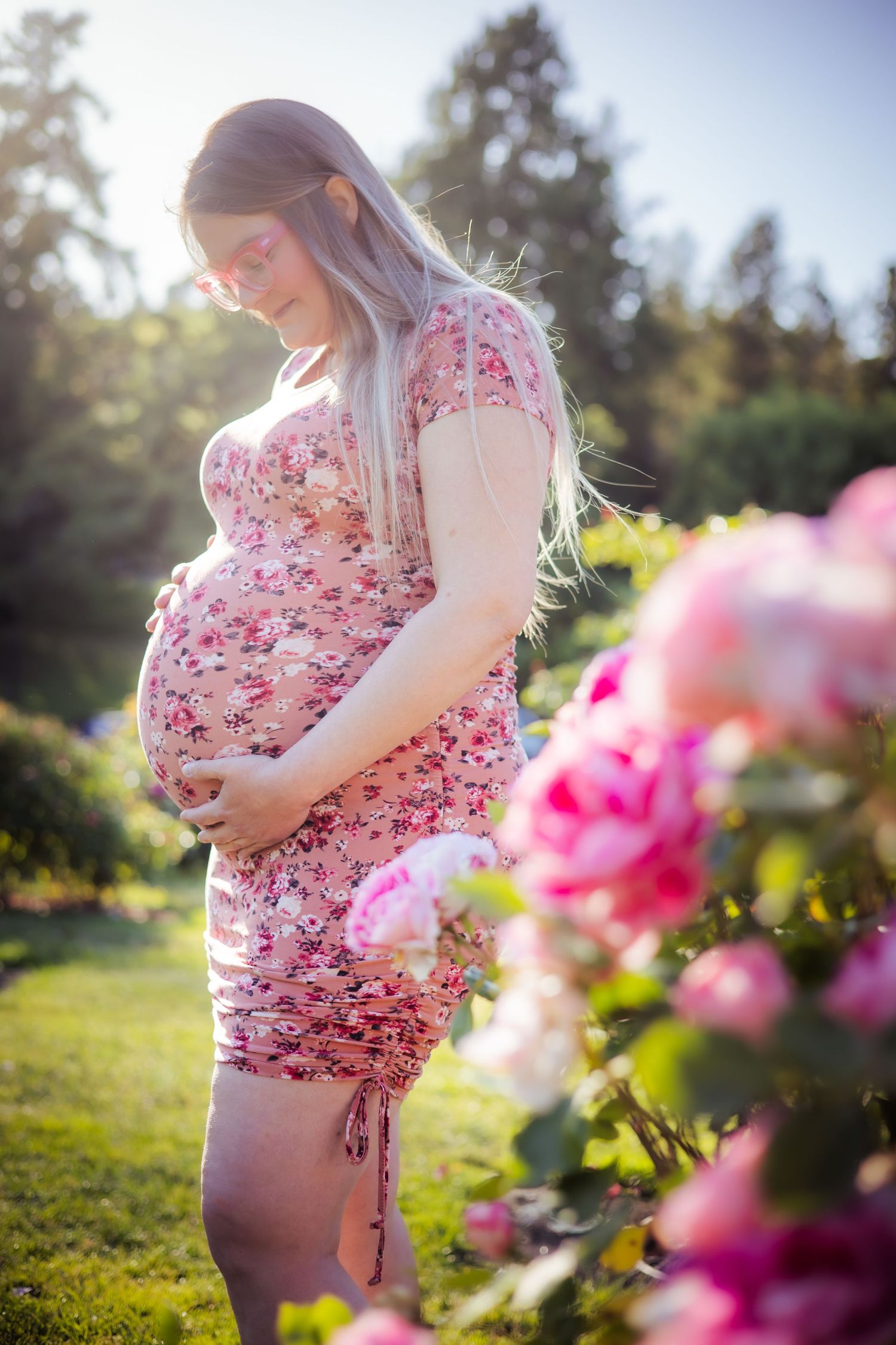 A pregnant woman in a floral dress stands in a garden with pink flowers during golden hour sunlight.