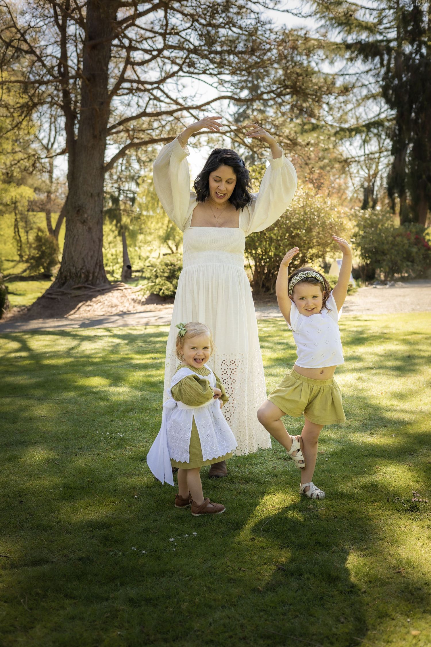 A joyful scene in a sunlit garden shows a woman in a white dress dancing with two small children on green grass.