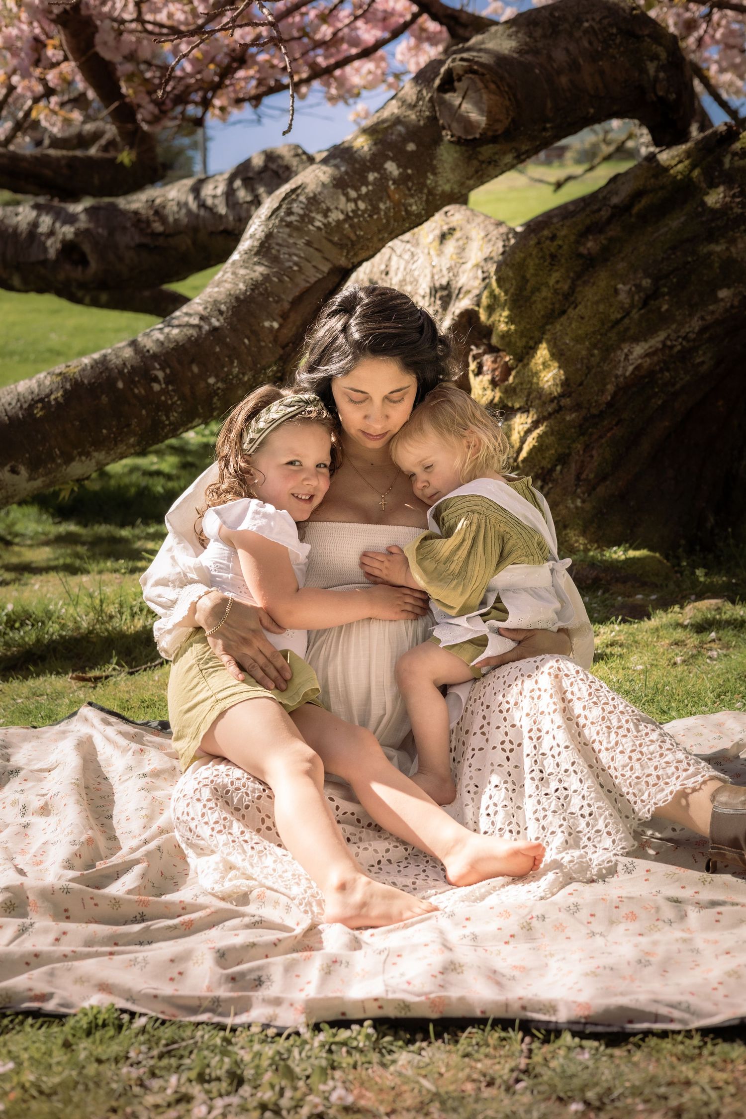 A tender family moment captured under a large tree on a picnic blanket in soft natural lighting.