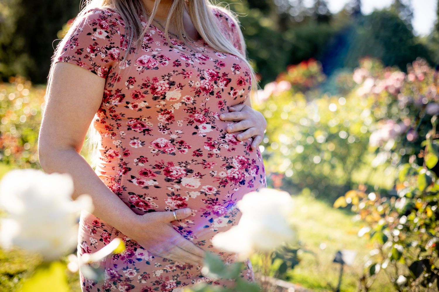 A person in a pink floral dress walks through a sunlit garden with blooming flowers and greenery.