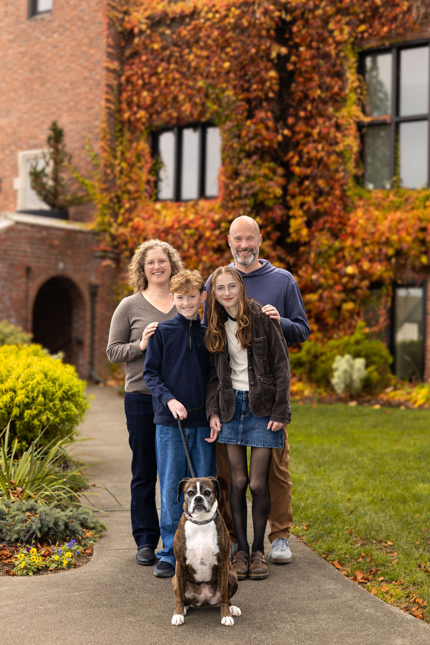 Family of four with a boxer dog posing in front of a brick building covered in autumn ivy foliage.