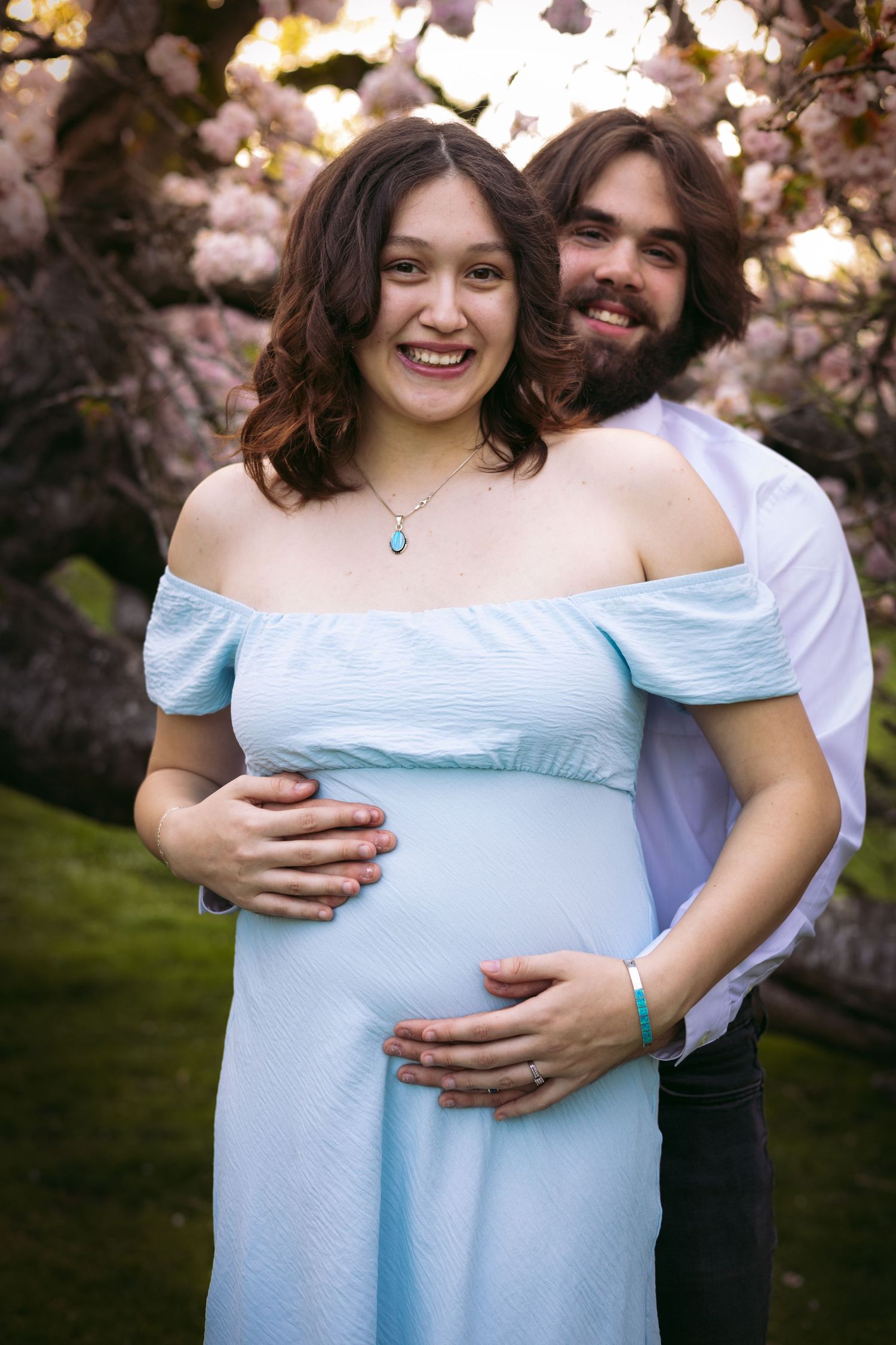 A couple embraces under blooming cherry blossoms, with the woman wearing a light blue off-shoulder maternity dress.