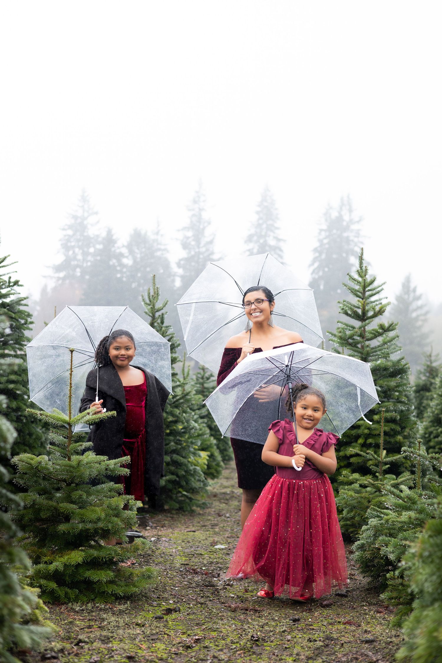 Family poses with umbrellas among evergreen trees at a Christmas tree farm on a misty day.