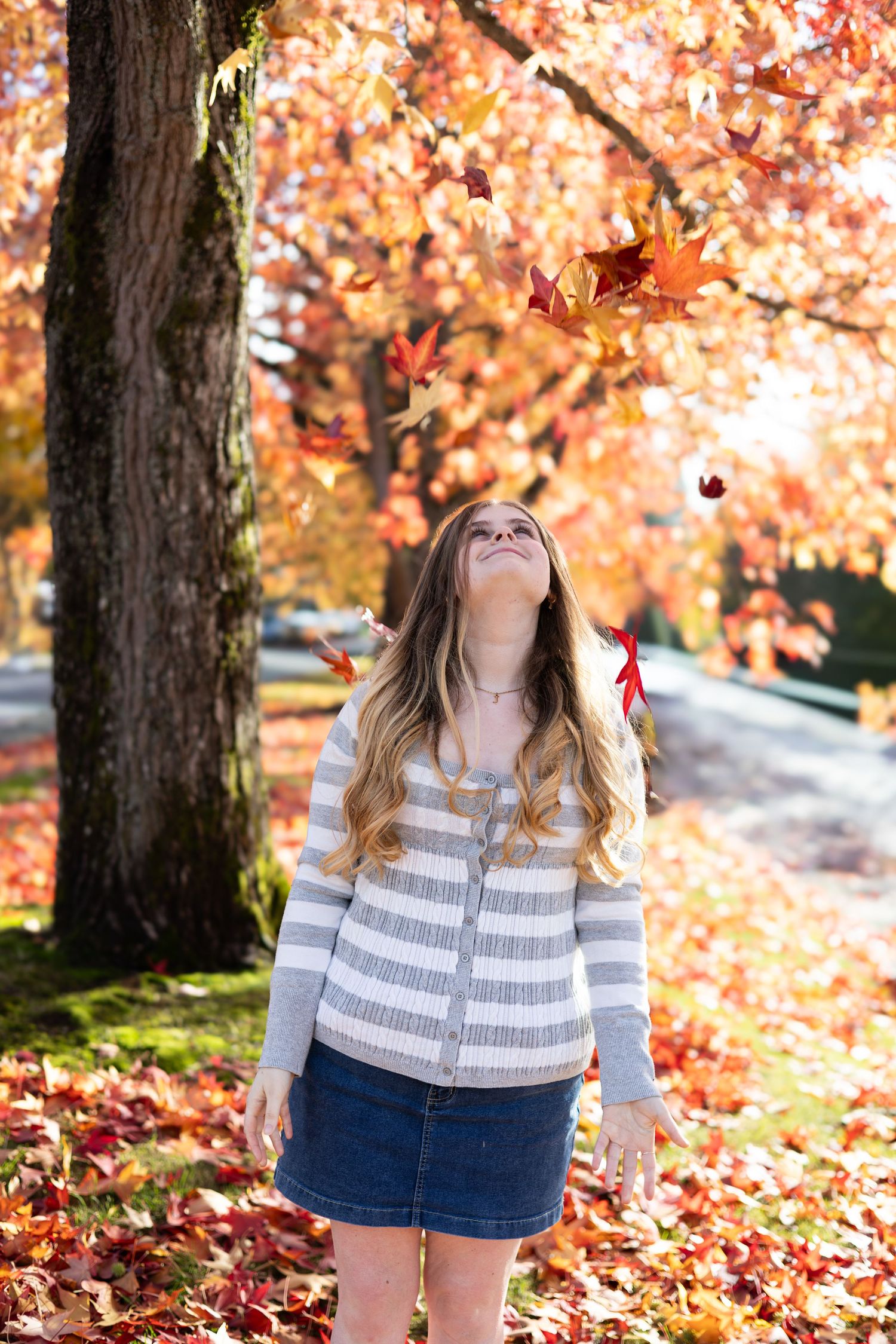 Young woman in striped top and denim skirt tossing colorful autumn leaves in the air outdoors.
