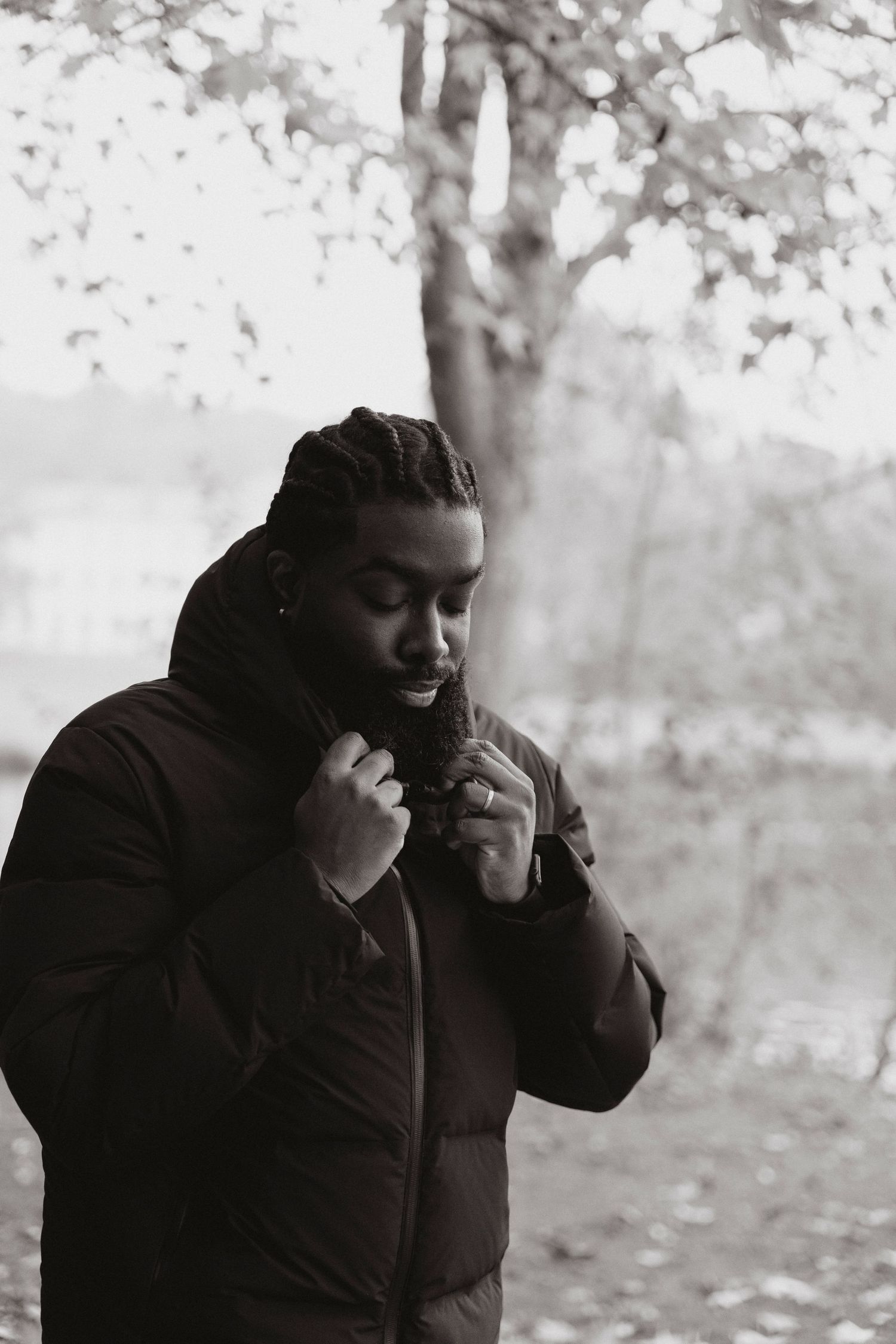 A black and white portrait of someone adjusting their winter coat in a park.