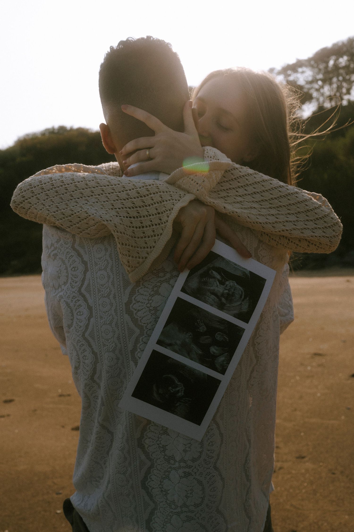 A couple embracing while holding ultrasound photos during sunset.
