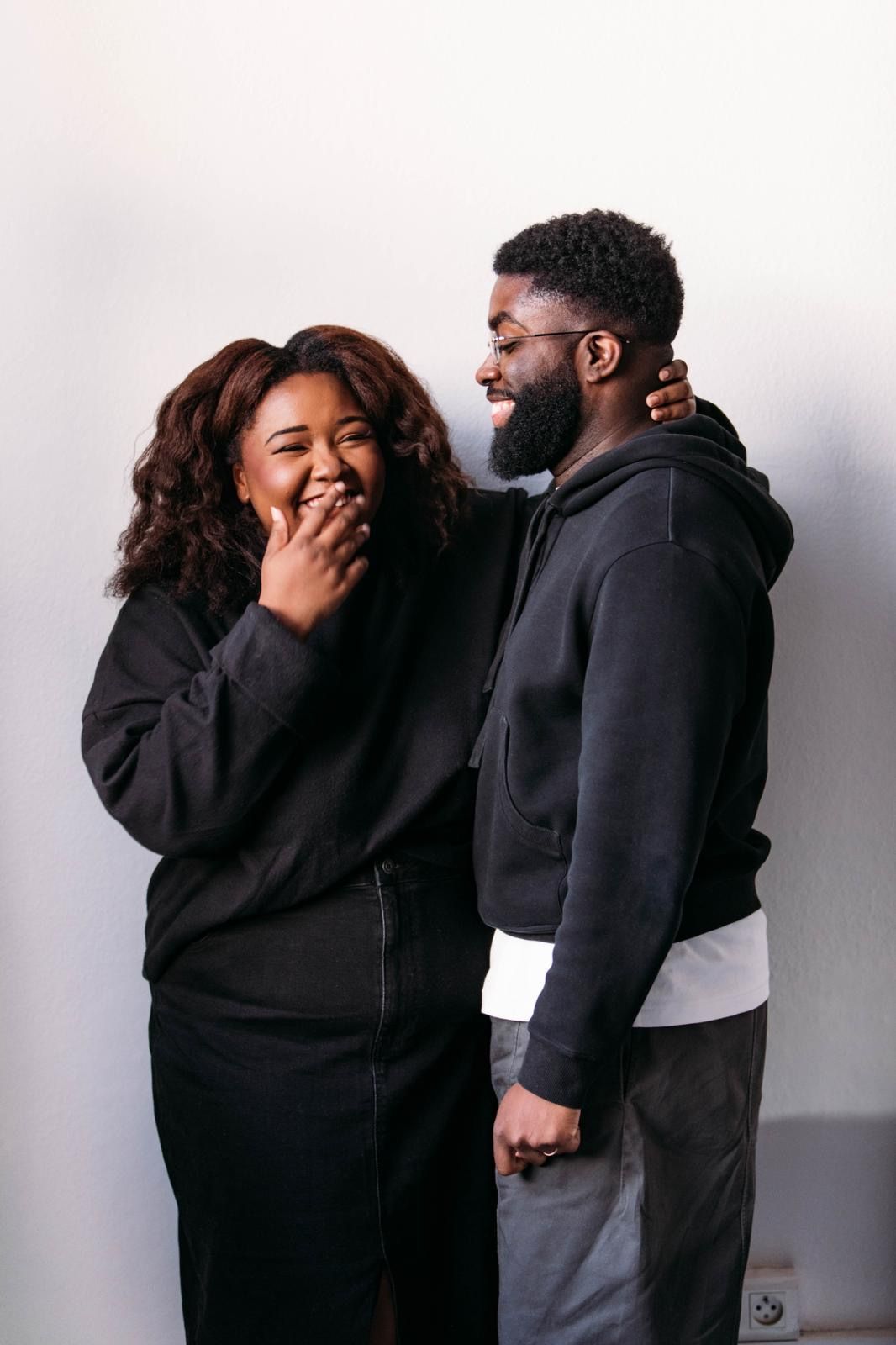 Candid moment of couple laughing together wearing matching black hoodies against white background.