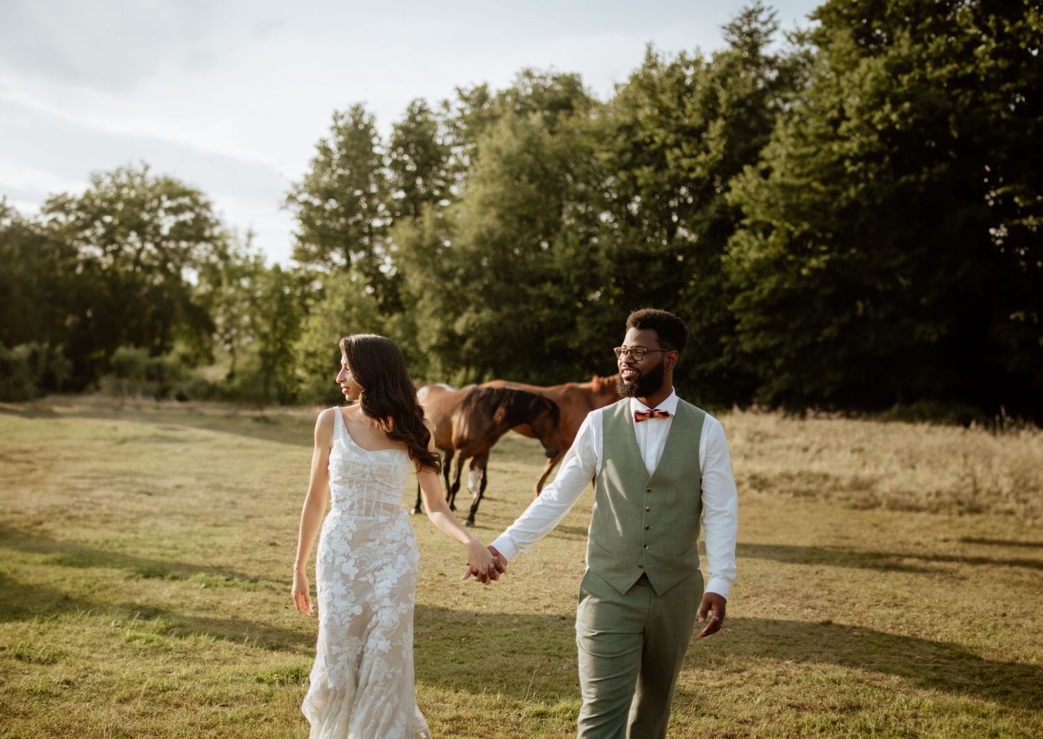 A couple in wedding attire walks hand in hand through a sunlit meadow with a horse grazing in the background.