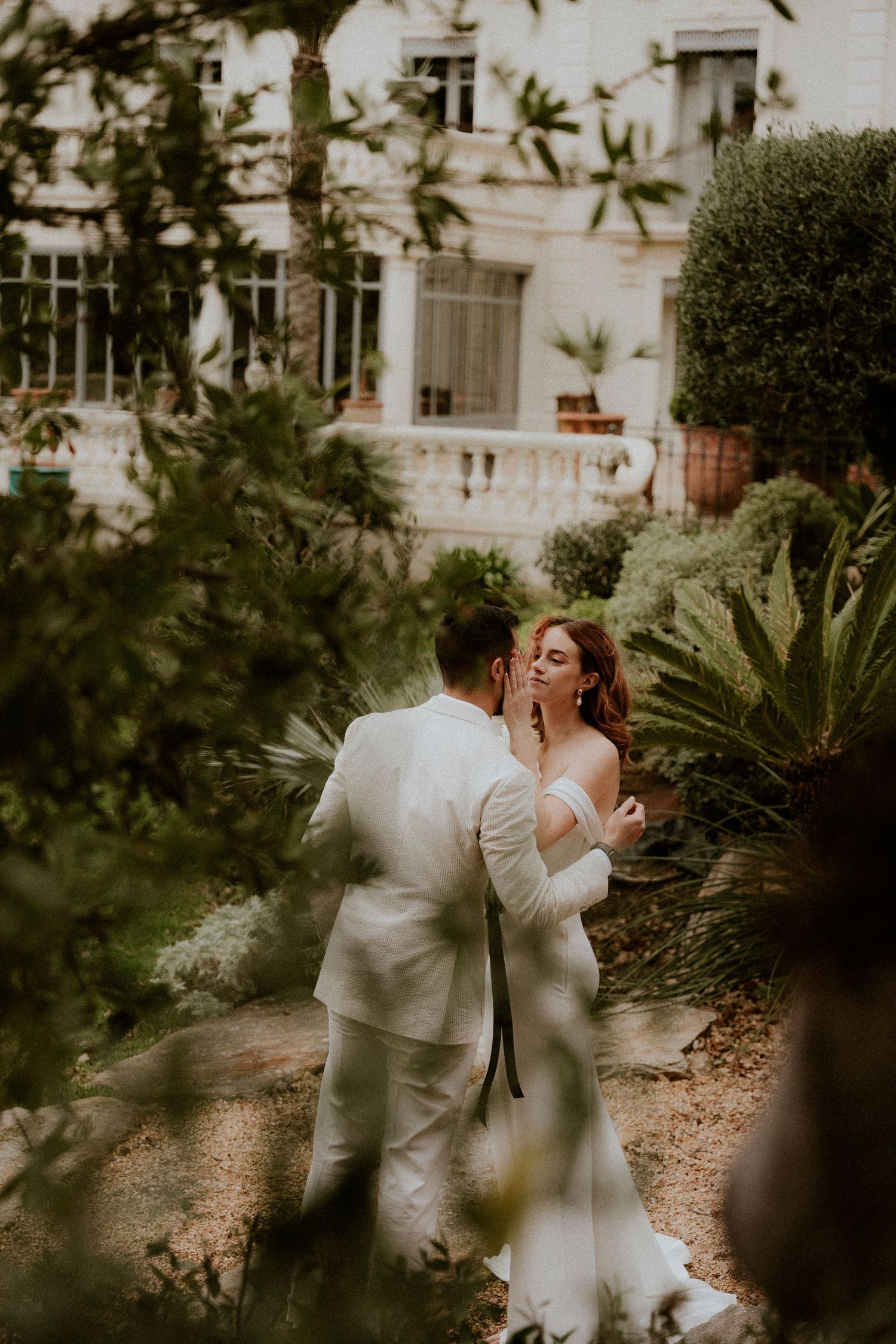 A romantic wedding moment captured through foliage near a historic white building.