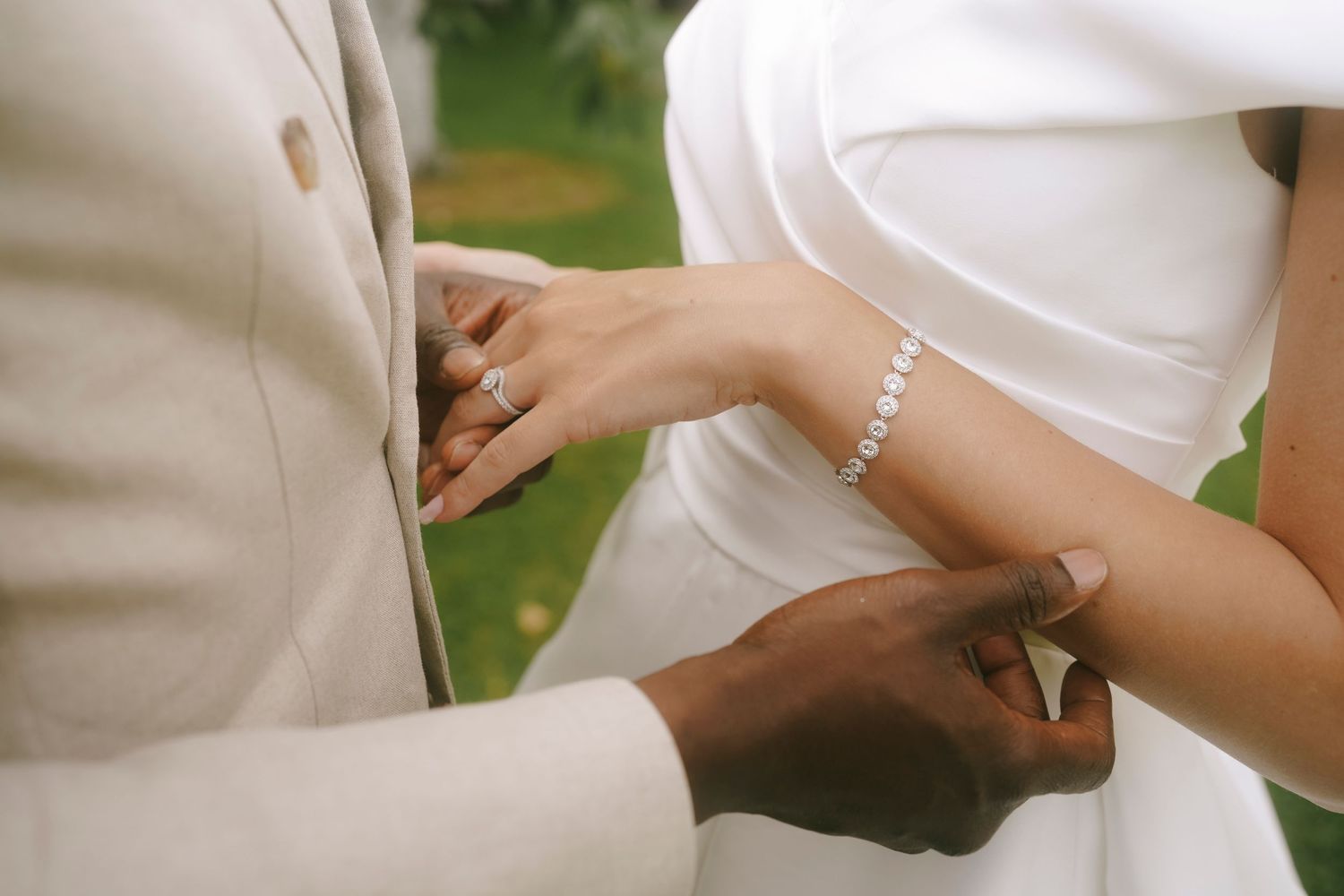 Two hands gently touch during a wedding ceremony, with sparkly bracelets and formal attire visible.