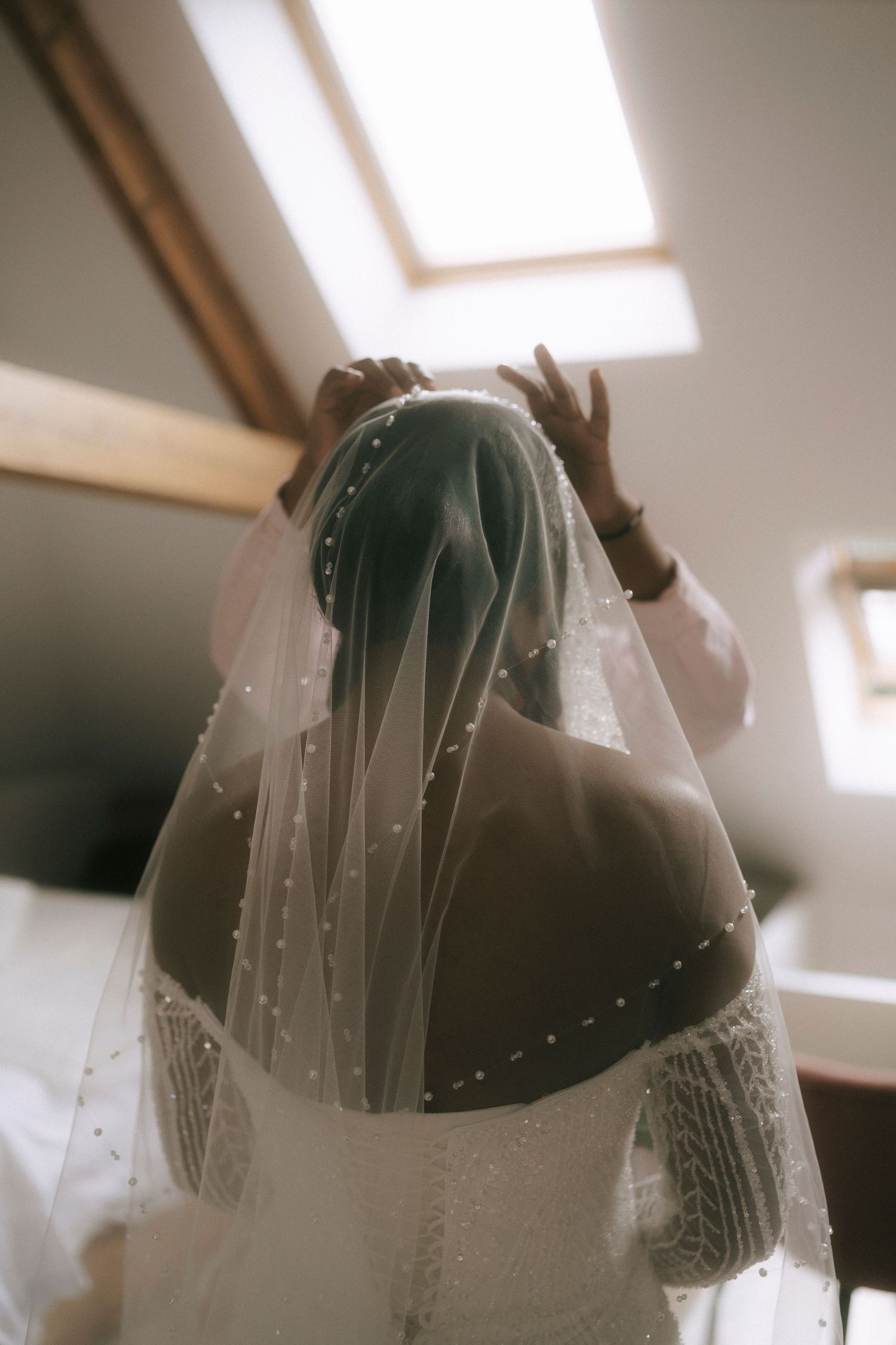 A bride wearing a delicate tulle veil with pearl details stands beneath a skylight in soft natural lighting.