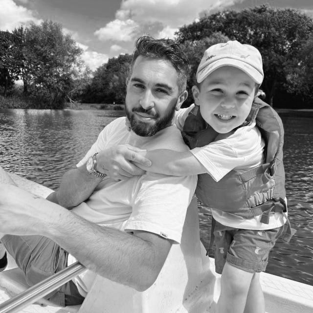 Man and young boy smiling on a boat near a river, black and white photo outdoors.