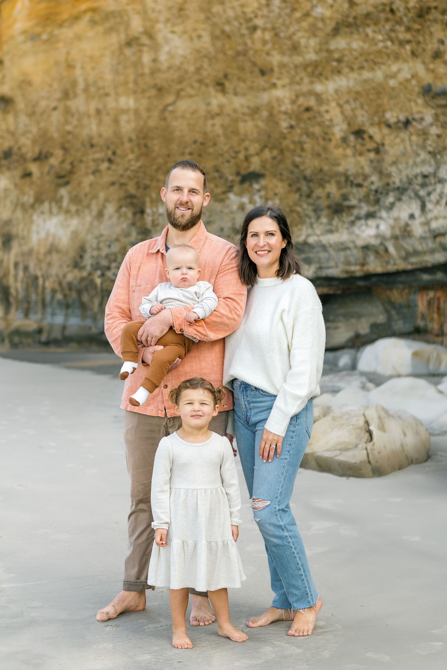 Barefoot family posing together on a sandy beach with rocky cliffs in the background.
