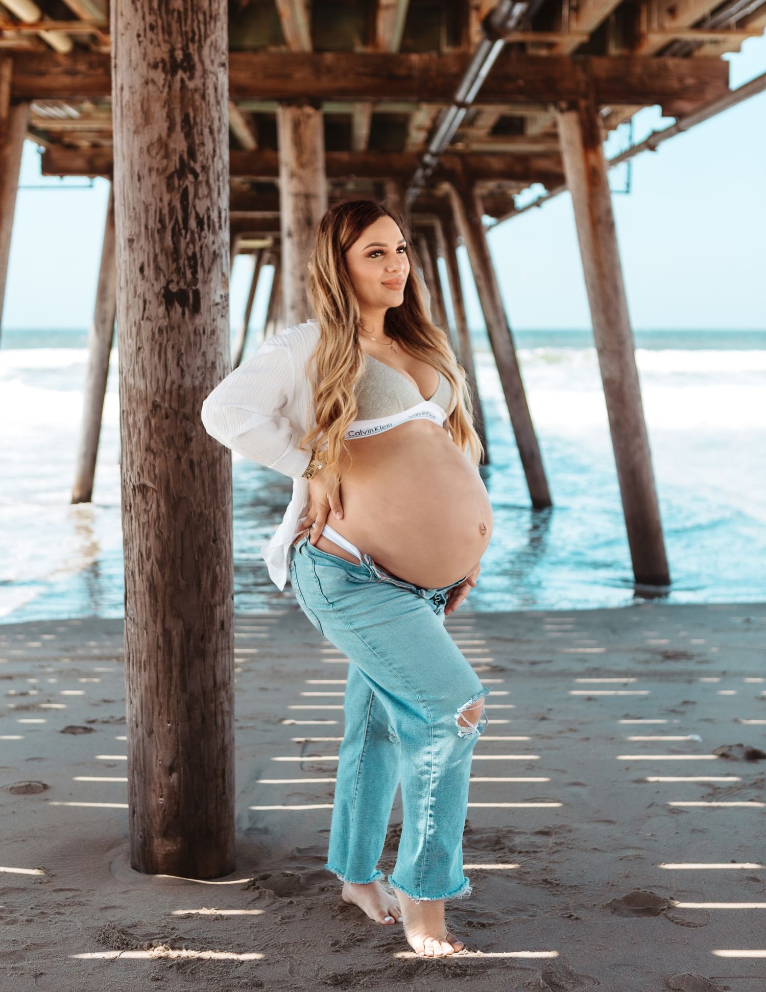 Pregnant woman poses under pier wearing ripped jeans and white top with ocean waves behind.